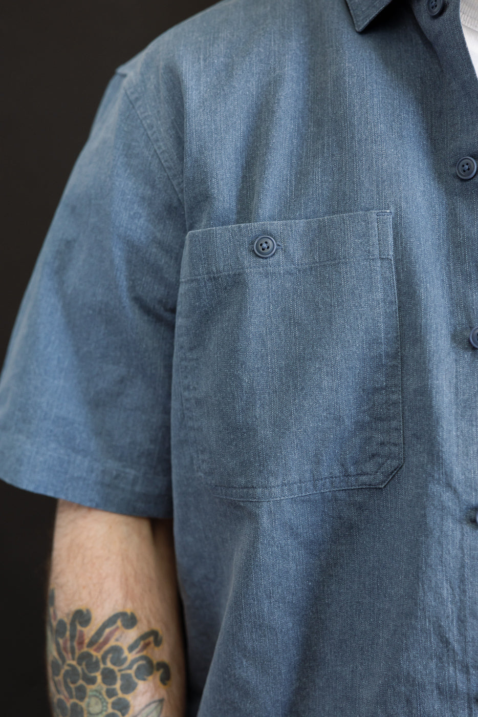 Close-up of the Short Sleeve Work Shirt - Blue Pigment Dye, showcasing its chambray-style fabric and square chest pocket, worn with a visible floral tattoo on the forearm and a hint of an undershirt.
