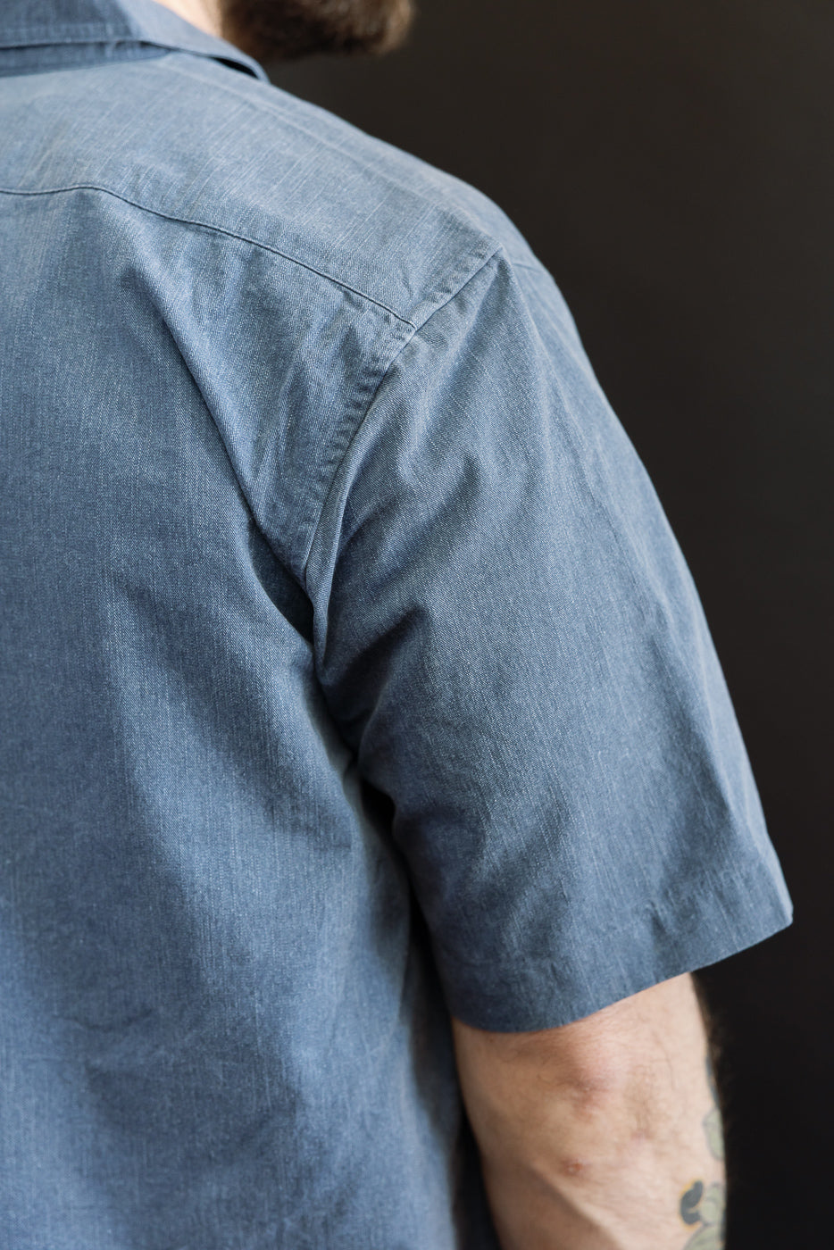 Close-up of a person wearing the 3sixteen Short Sleeve Work Shirt in blue pigment dye, showcasing the chambray-like fabric and short sleeves with a tattoo partially visible on the forearm.