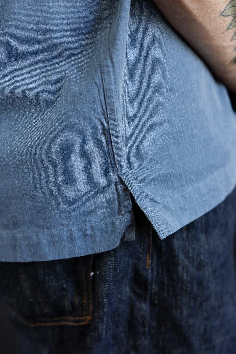 Close-up of the 3sixteen Short Sleeve Work Shirt - Blue Pigment Dye, showcasing its chambray fabric, untucked over dark-wash jeans, highlighting dual cut-corner chest pockets and Corozo buttons for a vintage look.