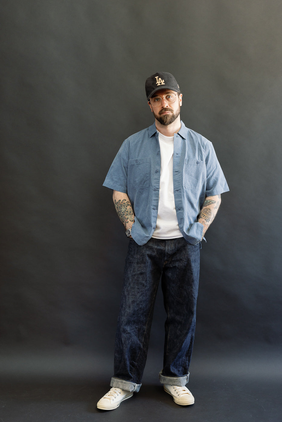 Man in Short Sleeve Work Shirt - Blue Pigment Dye, styled open over a white T-shirt, showcasing dual cut-corner chest pockets and Corozo buttons, paired with cuffed dark-denim jeans and sneakers.