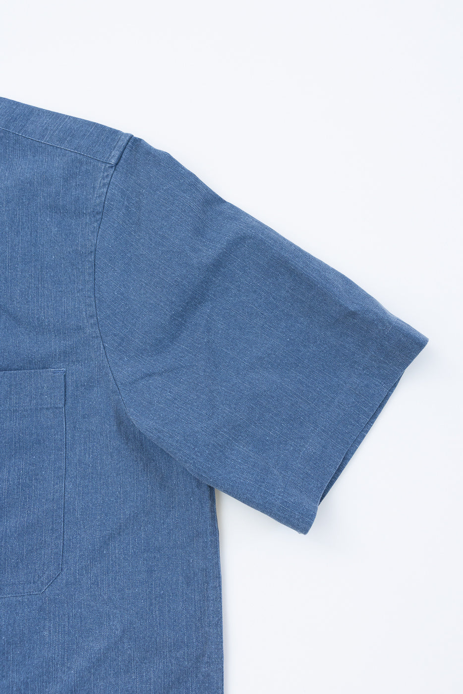 Close-up of the Short Sleeve Work Shirt - Blue Pigment Dye, featuring a chambray-textured fabric with a patch pocket and sleeve visible, showcasing its modern workwear design with dual cut-corner chest pockets.