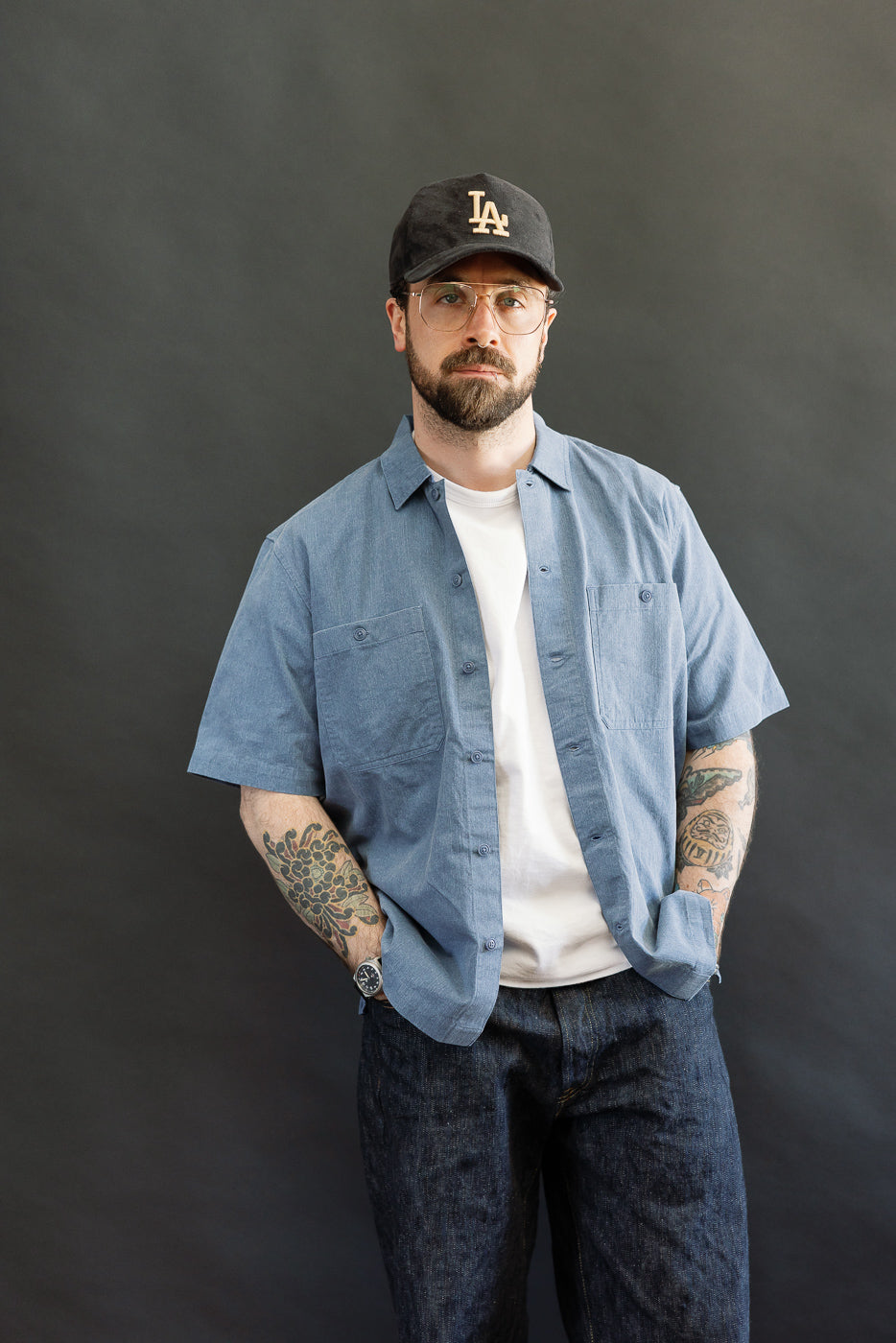 Man models the Short Sleeve Work Shirt - Blue Pigment Dye, showcasing its light blue, open short-sleeve design over a white T-shirt, featuring dual cut-corner chest pockets and Corozo button details.