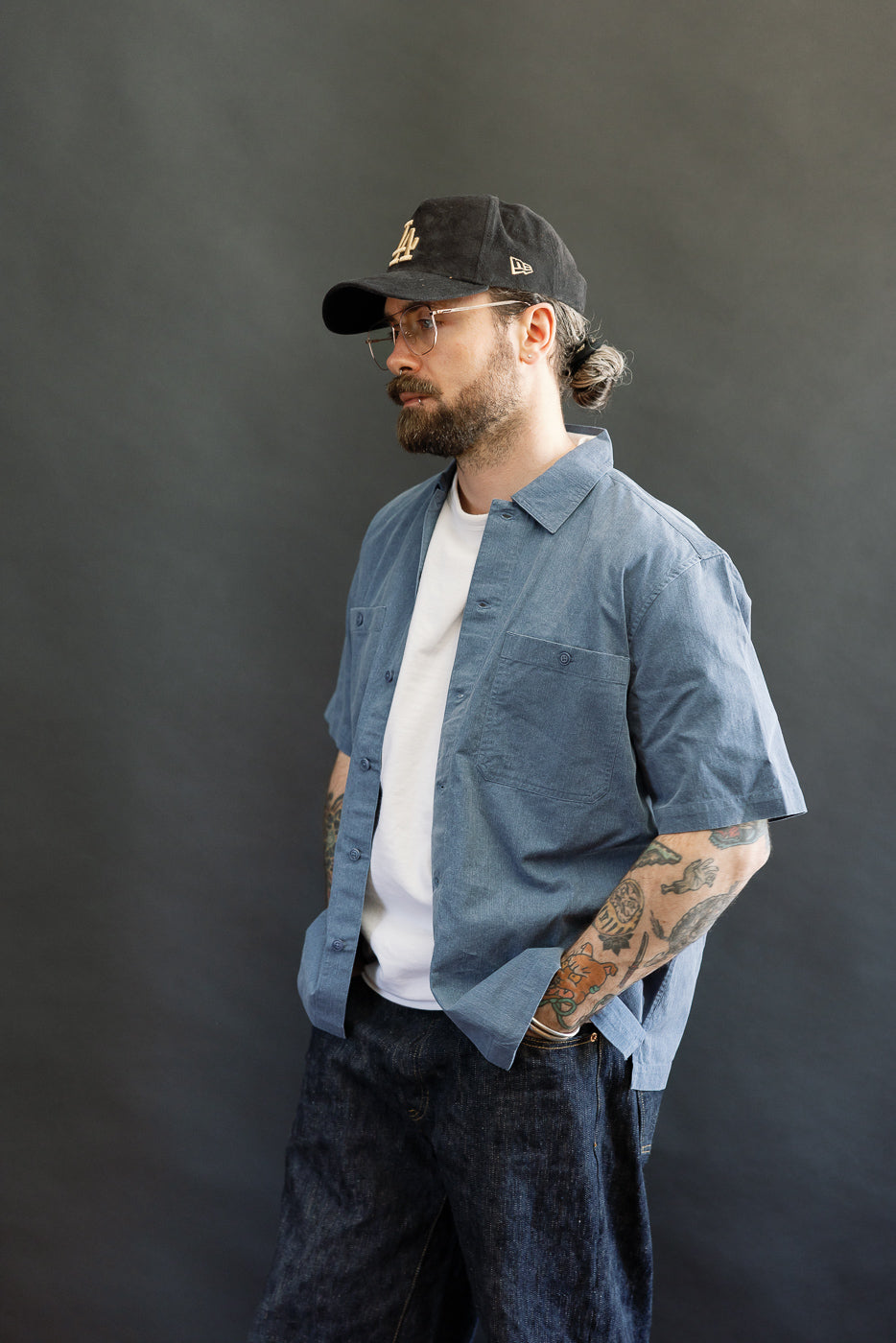 A casually dressed man models the 3sixteen Short Sleeve Work Shirt in blue pigment dye, layered over a white T-shirt. The shirt features dual cut-corner chest pockets, complementing his tattooed forearms.