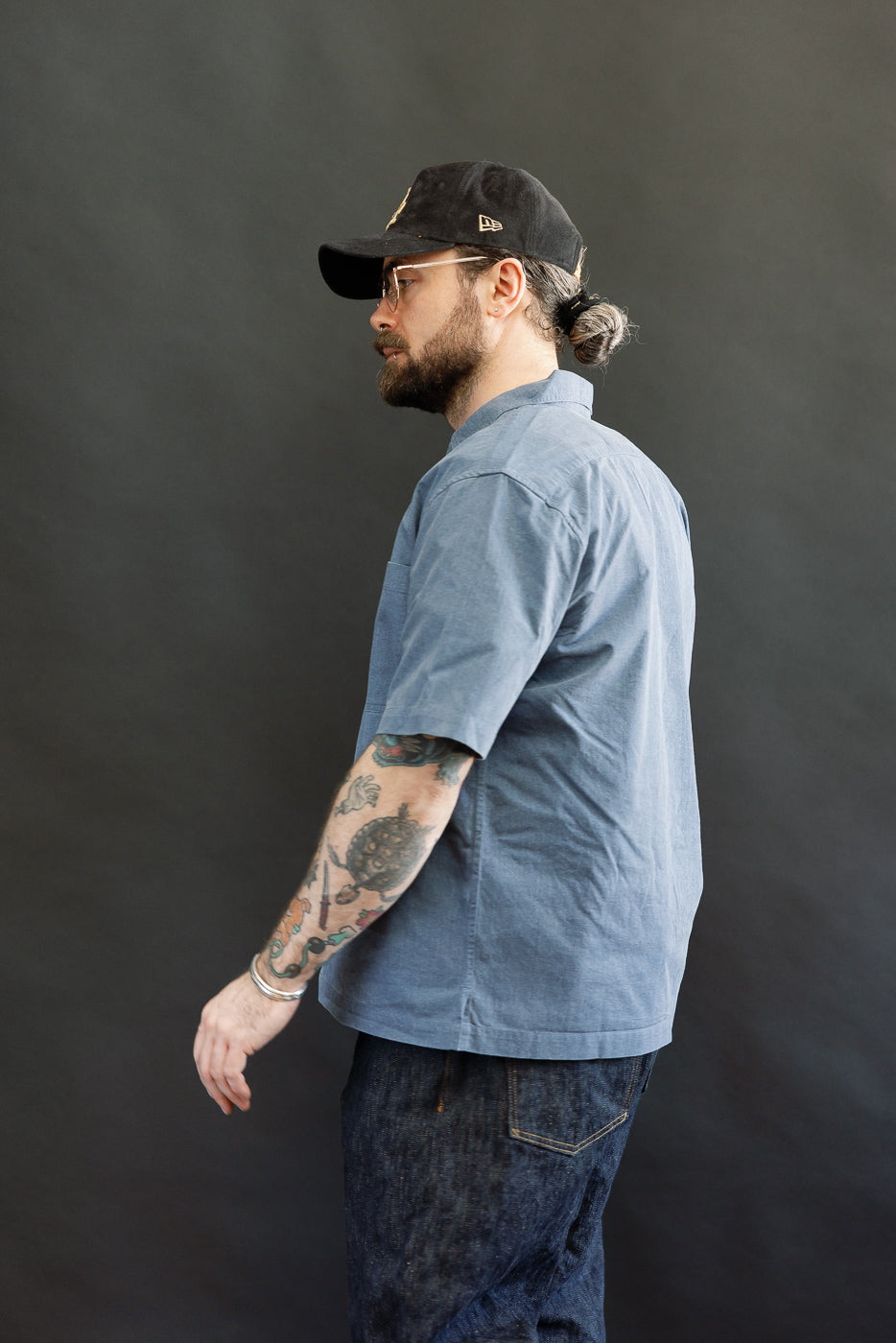 Man in profile wearing the Short Sleeve Work Shirt - Blue Pigment Dye with dual cut-corner chest pockets, clear-frame glasses, and black cap, showcasing its modern fit and vintage finish.