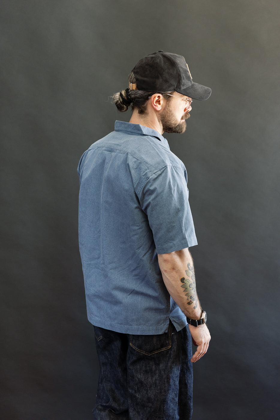 A man models the 3sixteen Short Sleeve Work Shirt in blue pigment dye, showcasing its generous, slightly cropped fit and dual cut-corner chest pockets, paired with dark jeans and a black baseball cap.