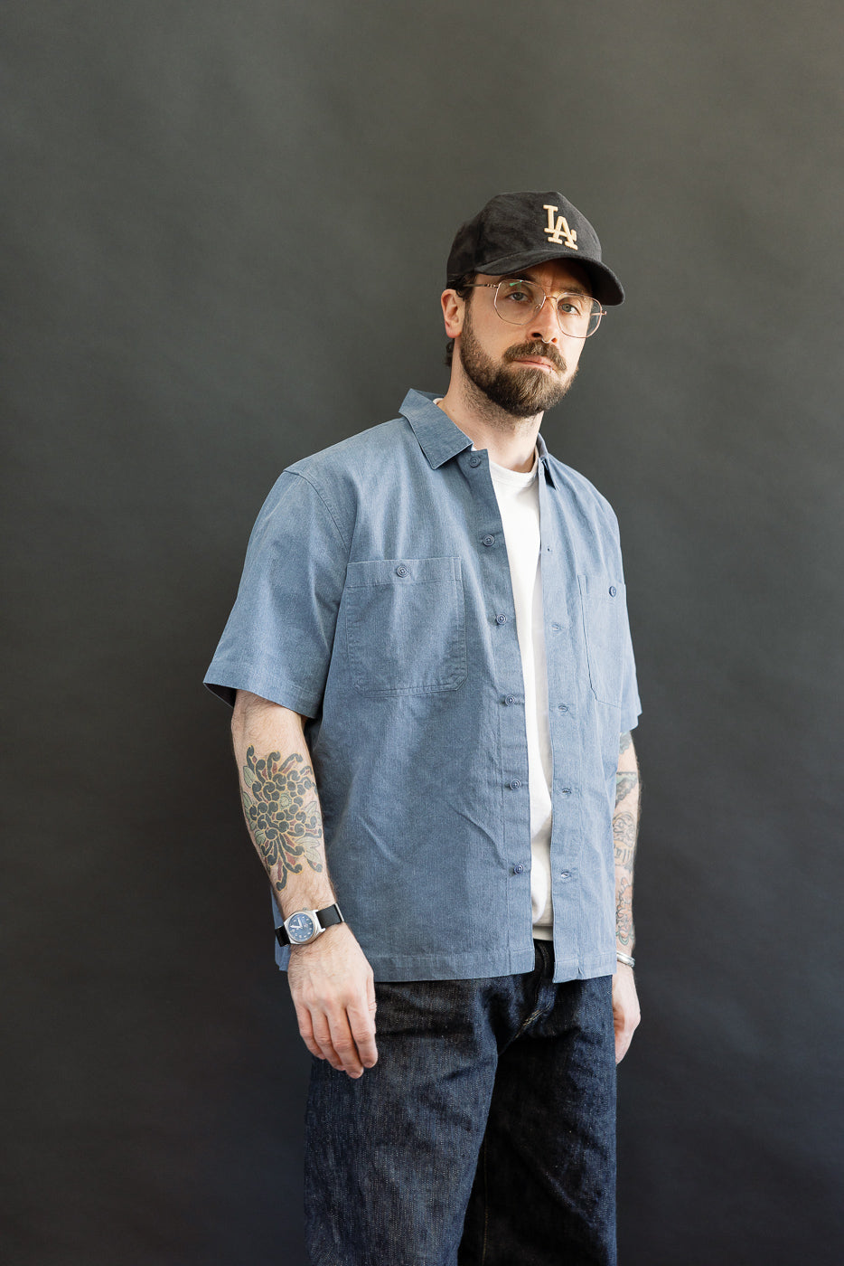 A bearded man models the Short Sleeve Work Shirt - Blue Pigment Dye, featuring dual chest pockets and Corozo buttons, layered over a white t-shirt, embodying a modern workwear aesthetic.