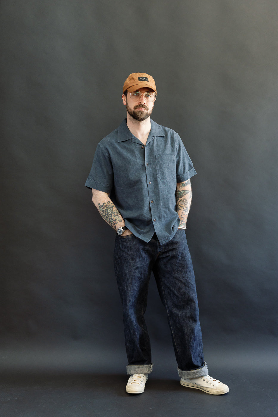 Full-length portrait of a man wearing the 3sixteen Vacation Shirt - Blue Linen with short sleeves, buttoned front, and an open collar, paired with dark denim jeans and off-white sneakers.