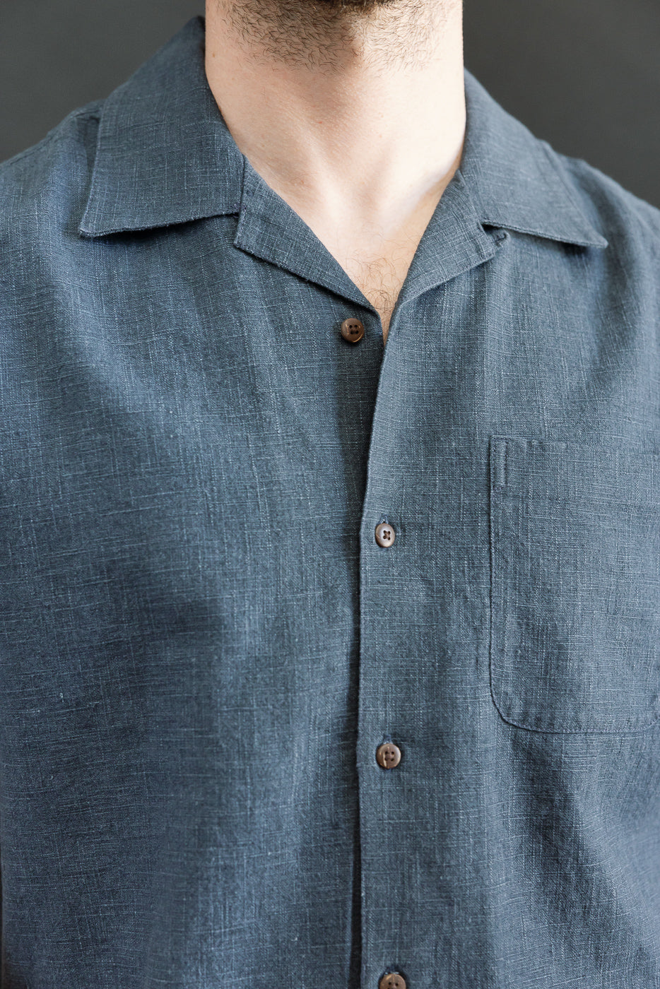 Person wearing the 3sixteen Vacation Shirt - Blue Linen, featuring a short-sleeve, camp-collar design with coconut buttons and a single chest pocket, showcasing a textured slate-blue fabric.