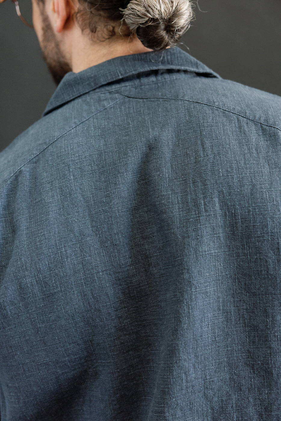 Close-up of the Vacation Shirt - Blue Linen's back, showcasing the textured linen fabric, open collar, and crossed yoke seam. The model's hair is styled in a small bun.