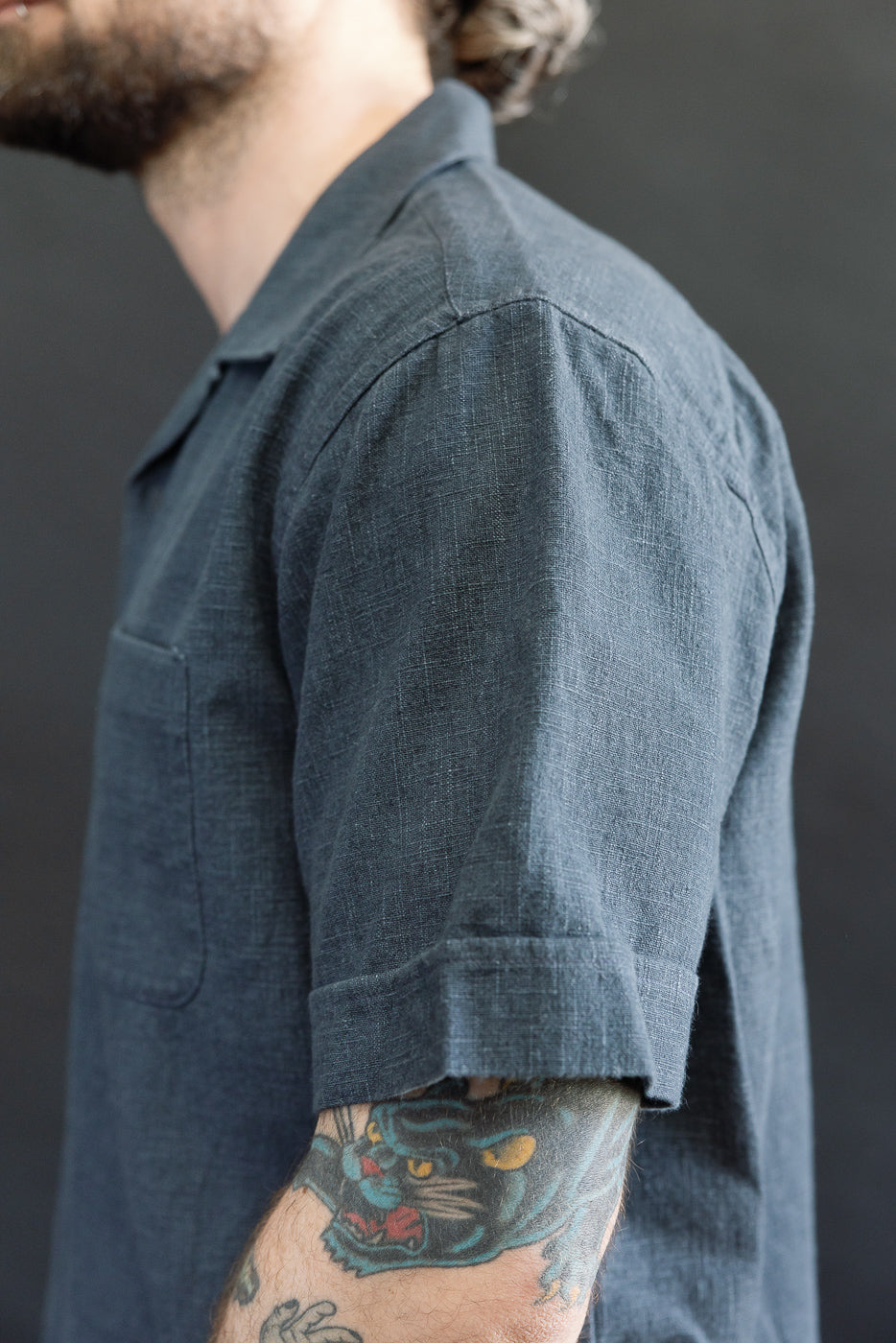 Close-up of the Vacation Shirt - Blue Linen on a model, showcasing a textured, short-sleeve button-up with a camp collar and chest pocket. The sleeve is lightly rolled, revealing a colorful tattoo.