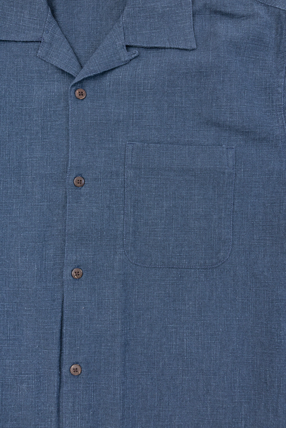 Close-up of the Vacation Shirt - Blue Linen, featuring a folded camp collar, chest patch pocket, and a row of four brown coconut buttons, showcasing its woven slub texture and relaxed design.