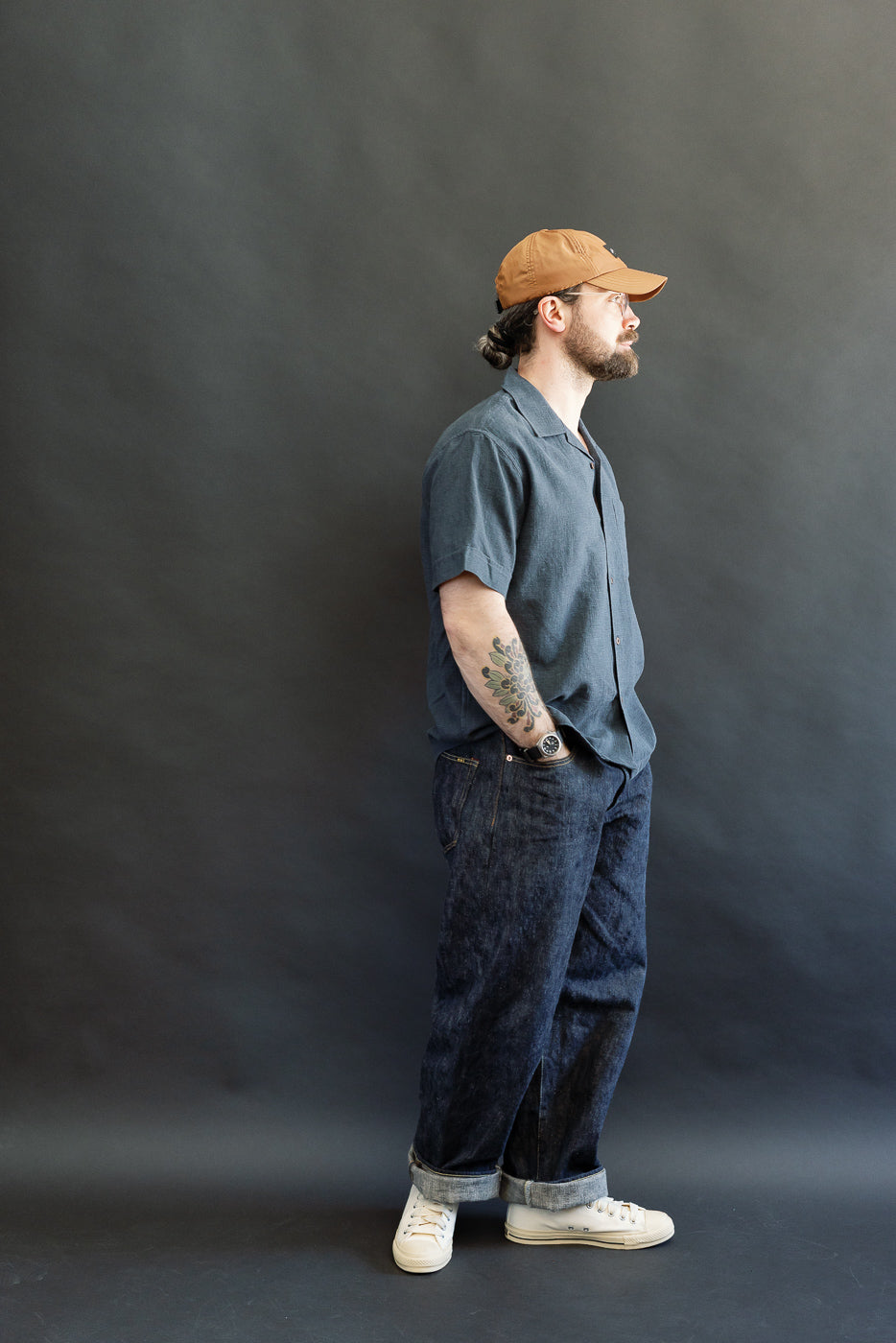 Man wearing the Vacation Shirt - Blue Linen, styled with dark denim jeans and white sneakers. The short-sleeve, open-collar shirt features a relaxed fit, coconut buttons, and a single chest pocket.