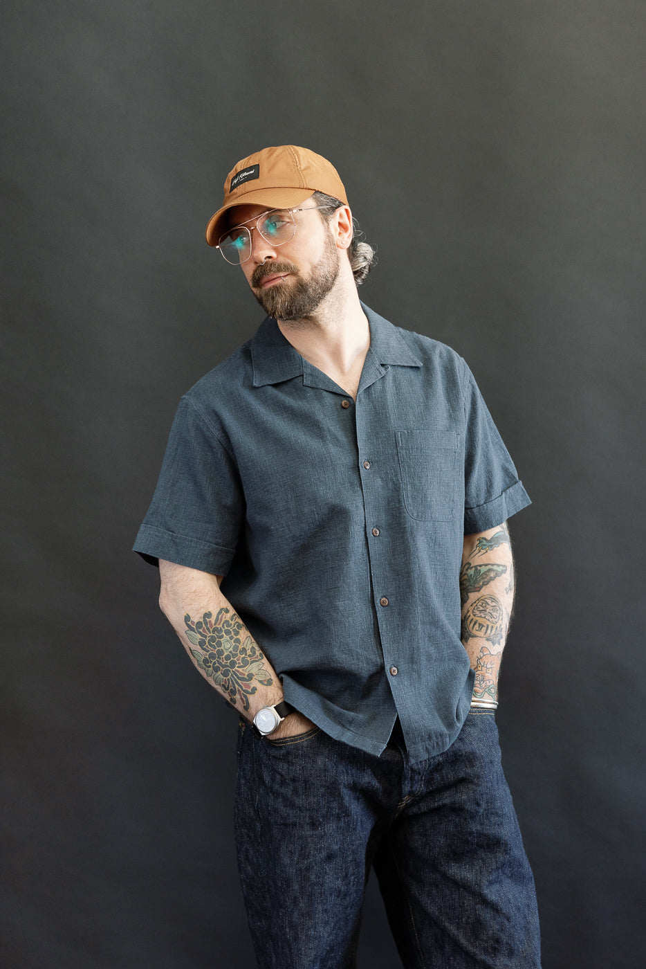 A bearded man models the 3sixteen Vacation Shirt - Blue Linen, showcasing its relaxed fit, open collar, and single chest pocket, with colorful tattoos visible on his forearms.