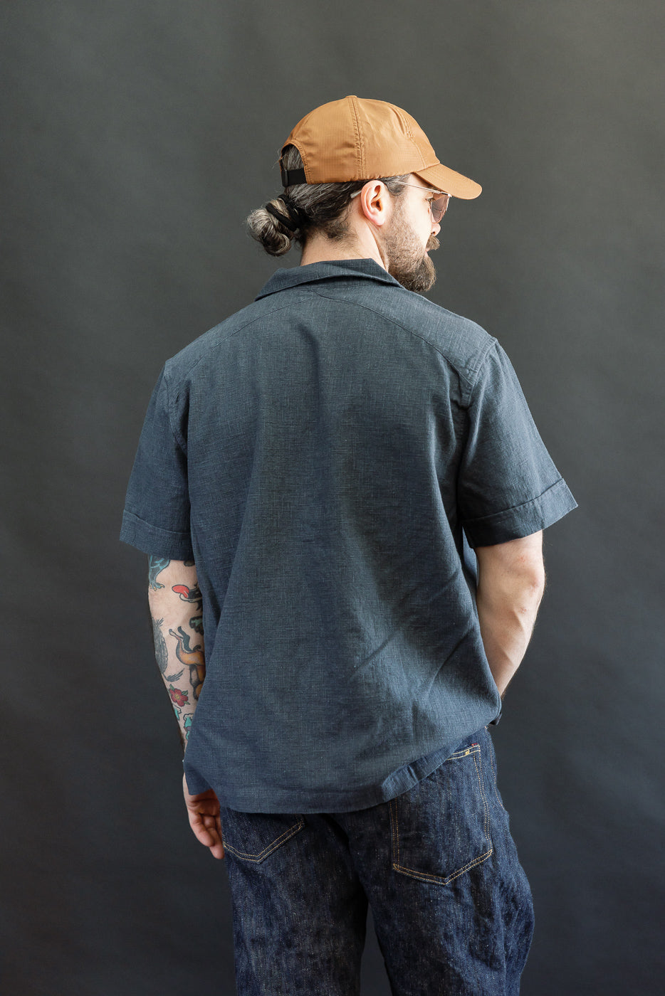 A man models the 3sixteen Vacation Shirt - Blue Linen, featuring a relaxed fit and open collar, styled casually with denim jeans and a backwards baseball cap against a dark backdrop.