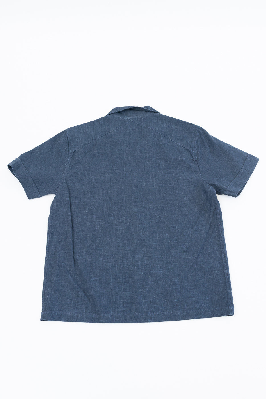 Vacation Shirt - Blue Linen, showcasing the back of a short-sleeved, deep blue, button-down shirt with a crossed-back yoke and coconut buttons, laid flat against a plain backdrop.