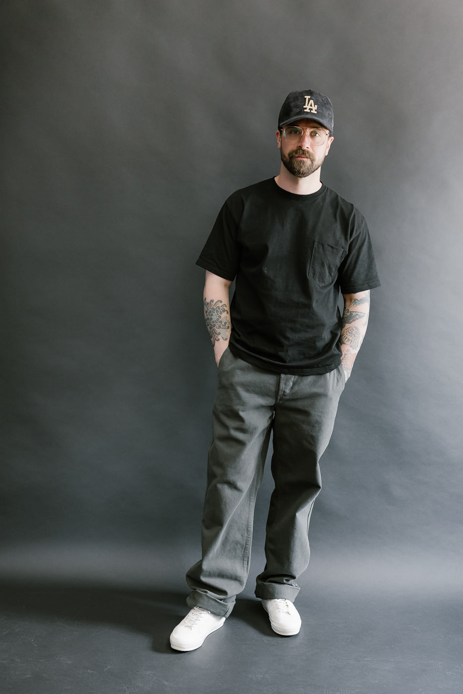 Man wearing a black t-shirt, gray pants, and a black cap against a gray background