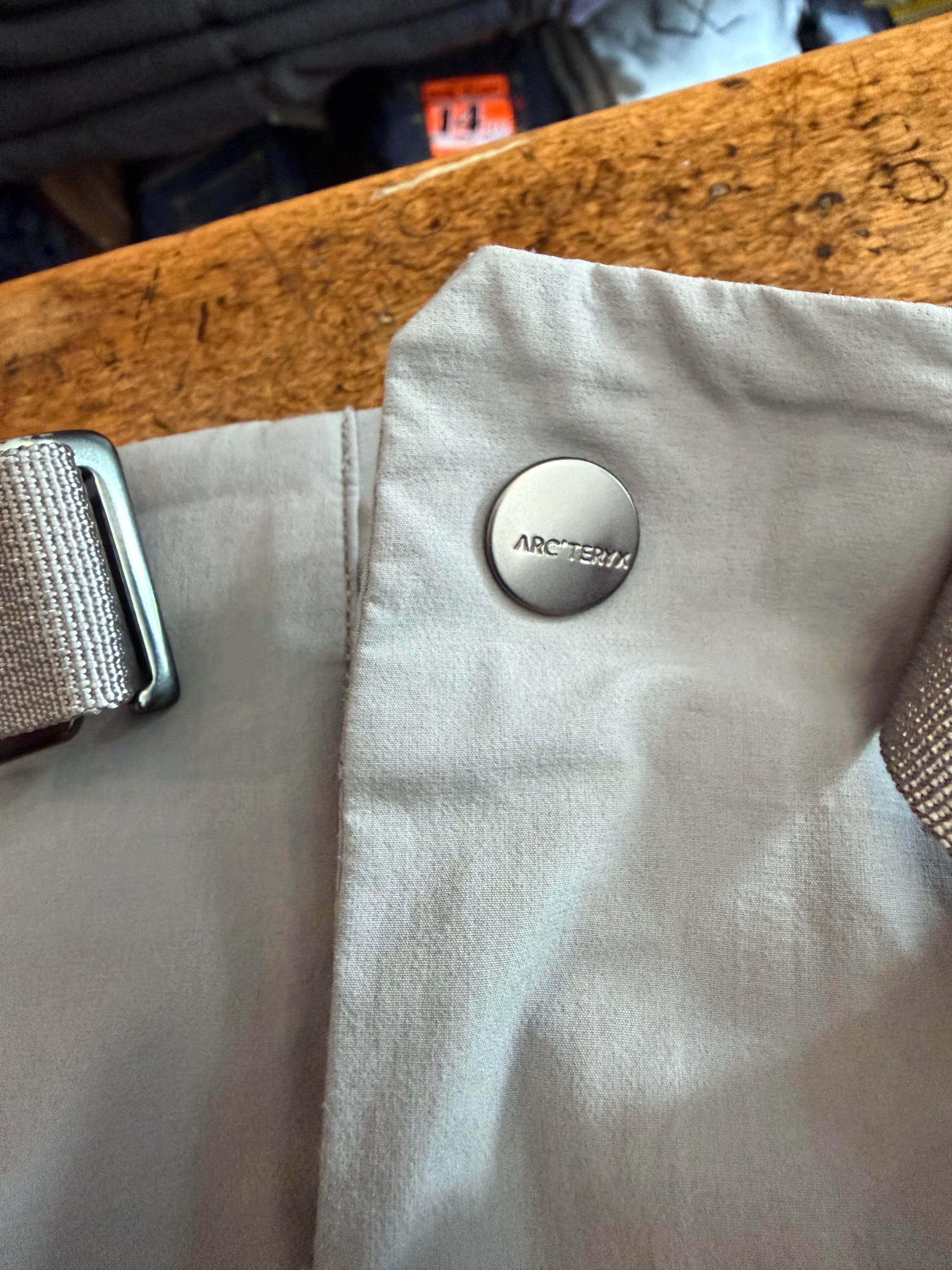 Close-up of Arc'teryx button on light gray garment front, showing fabric texture and metal buckle detail