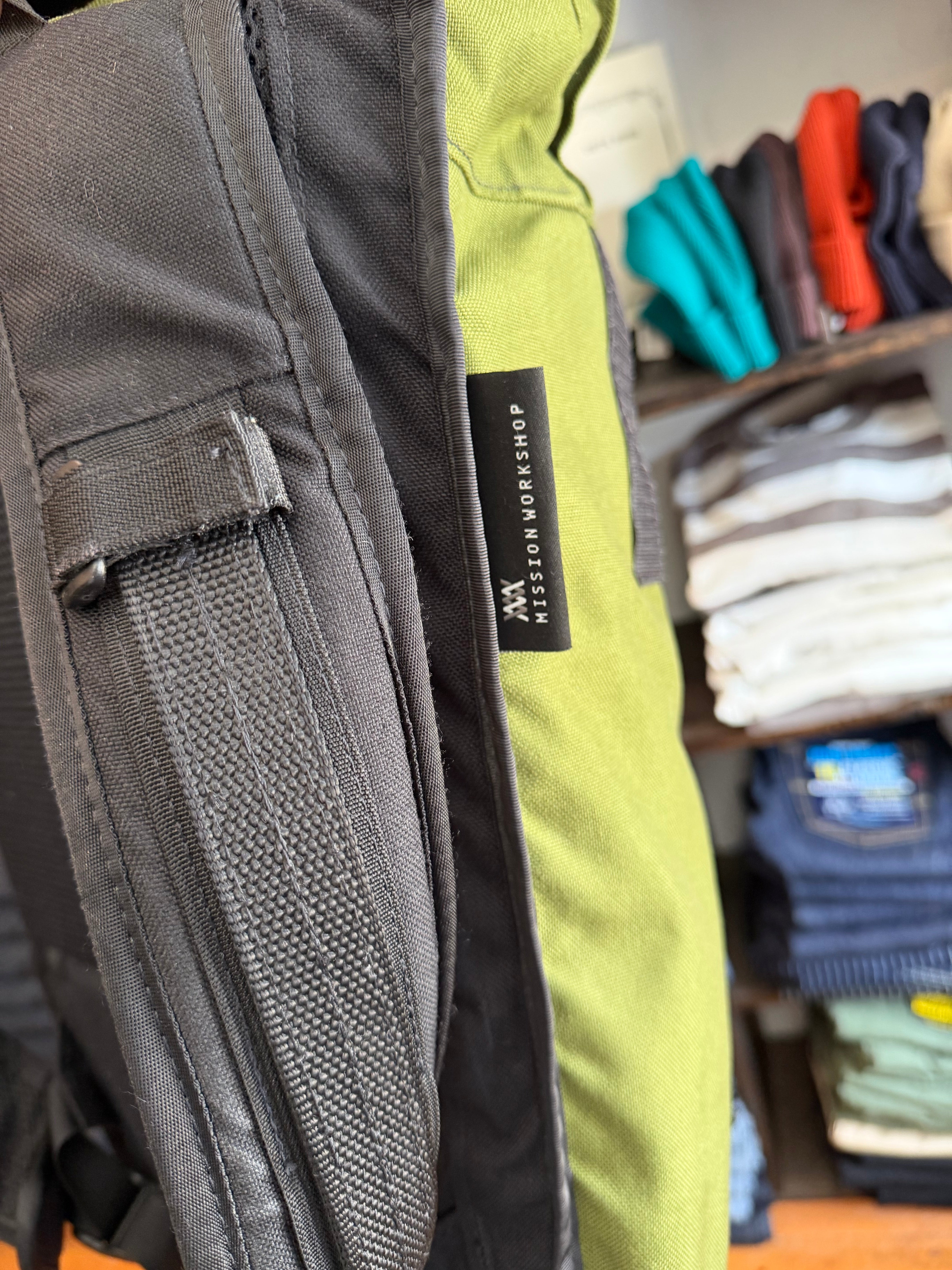 Close-up of gray cargo pants side pocket with button, green pants showing Mission Workshop tag, front view