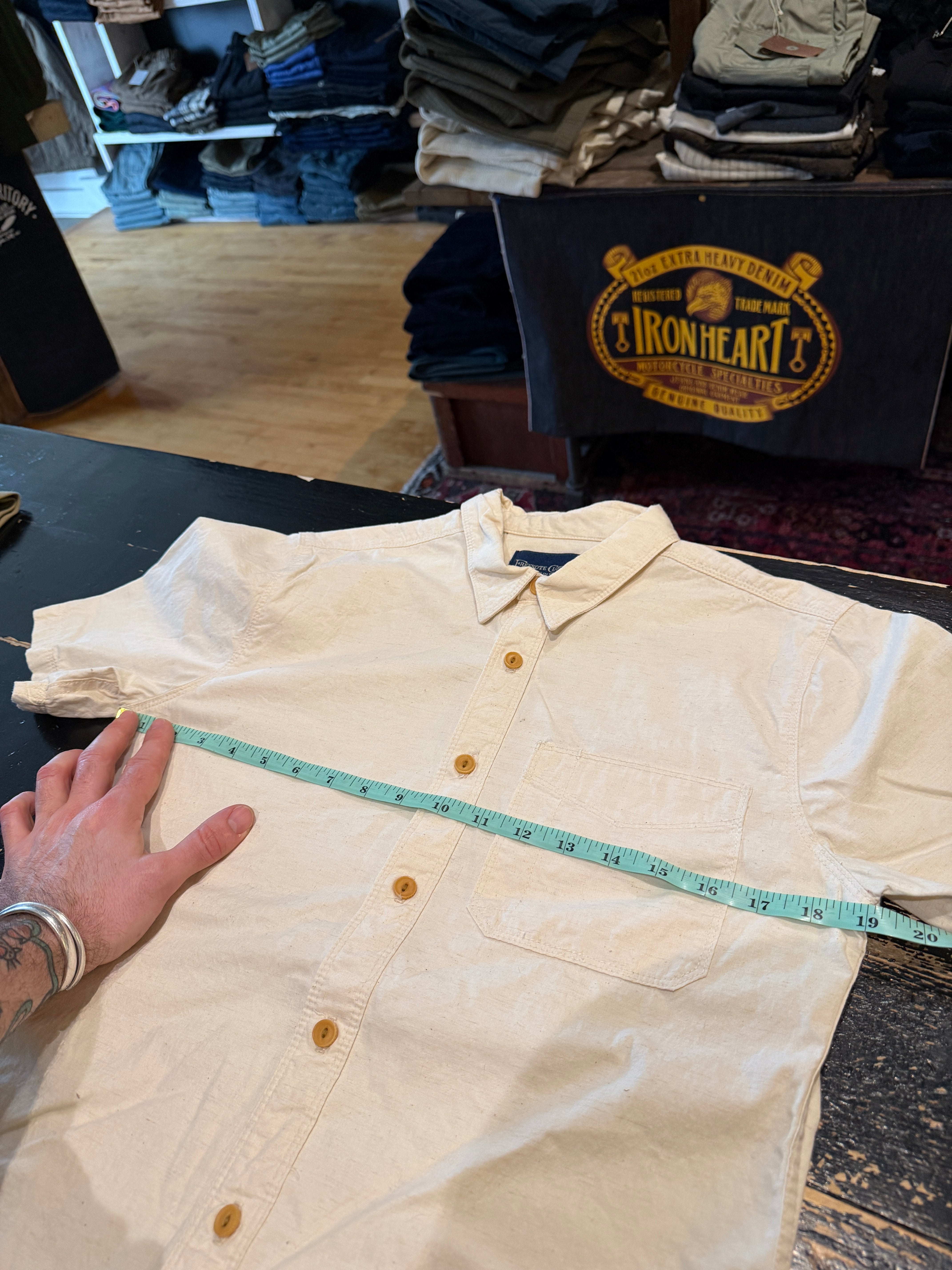 Front flat lay of cream short sleeve button-up shirt with chest pocket, being measured across chest with tape measure.