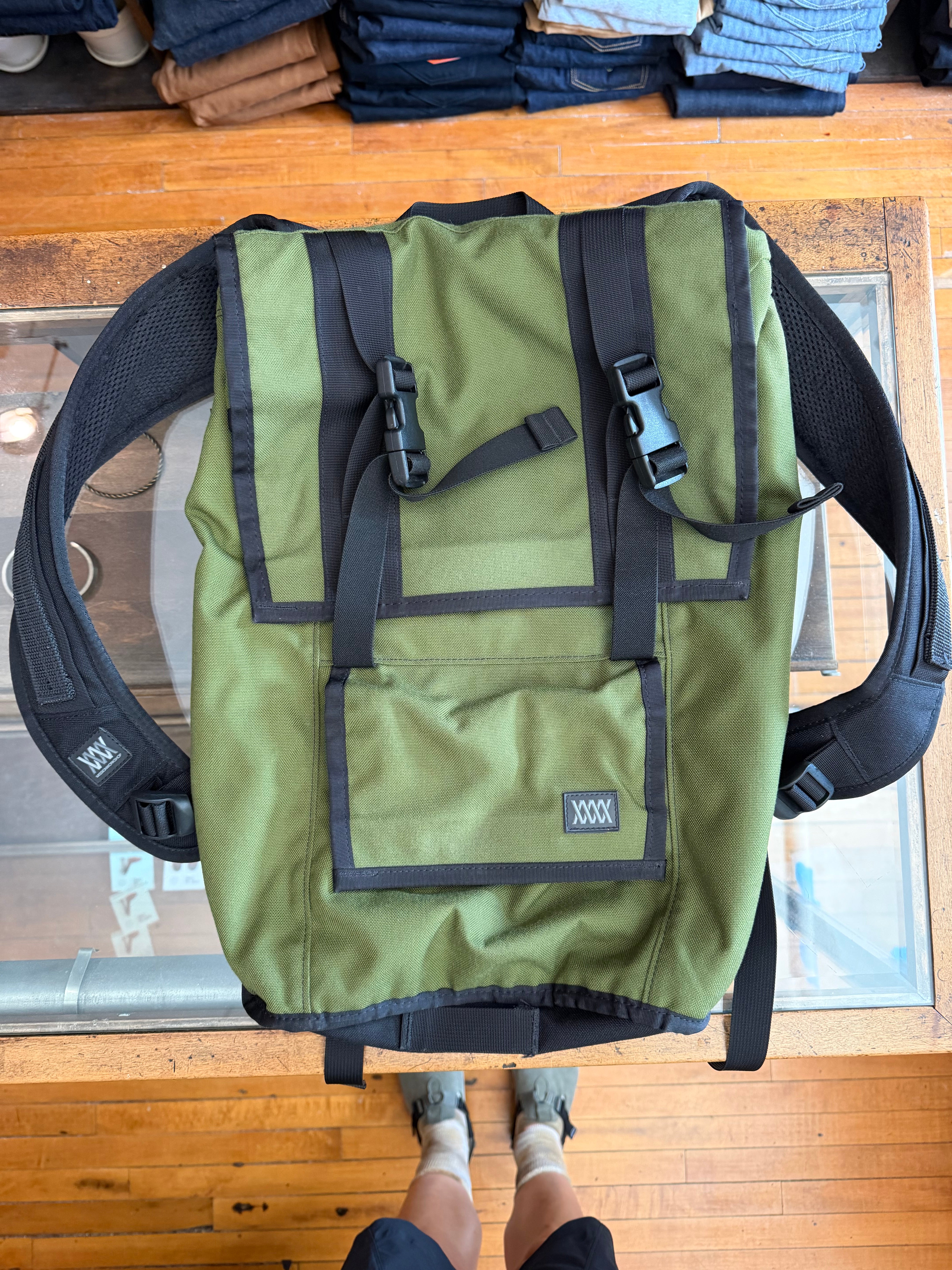 Olive green backpack with black straps, front view, showing buckles and brand tag, on glass table