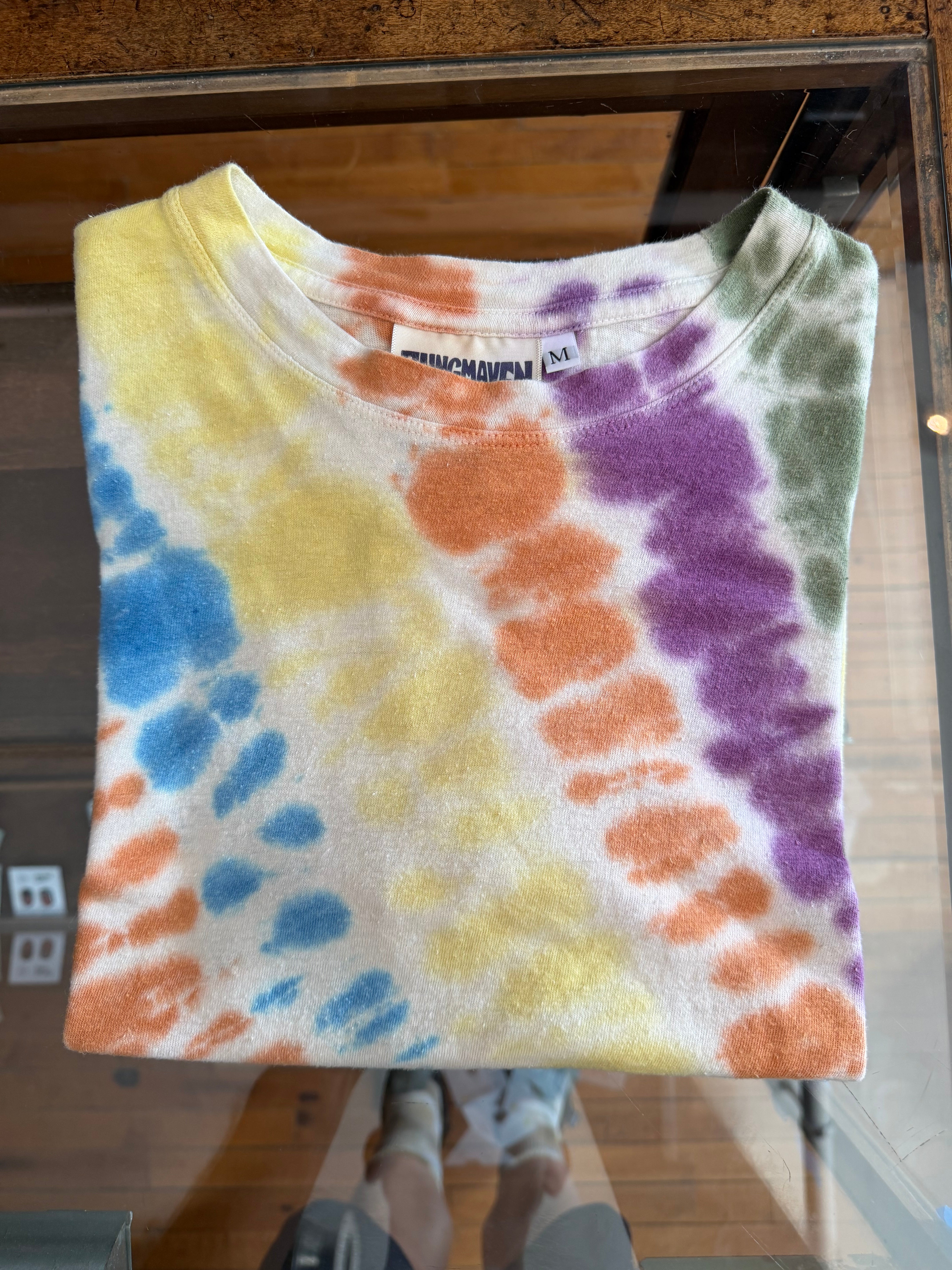Folded tie-dye t-shirt front view, multi-color spots, crew neck, size M tag visible