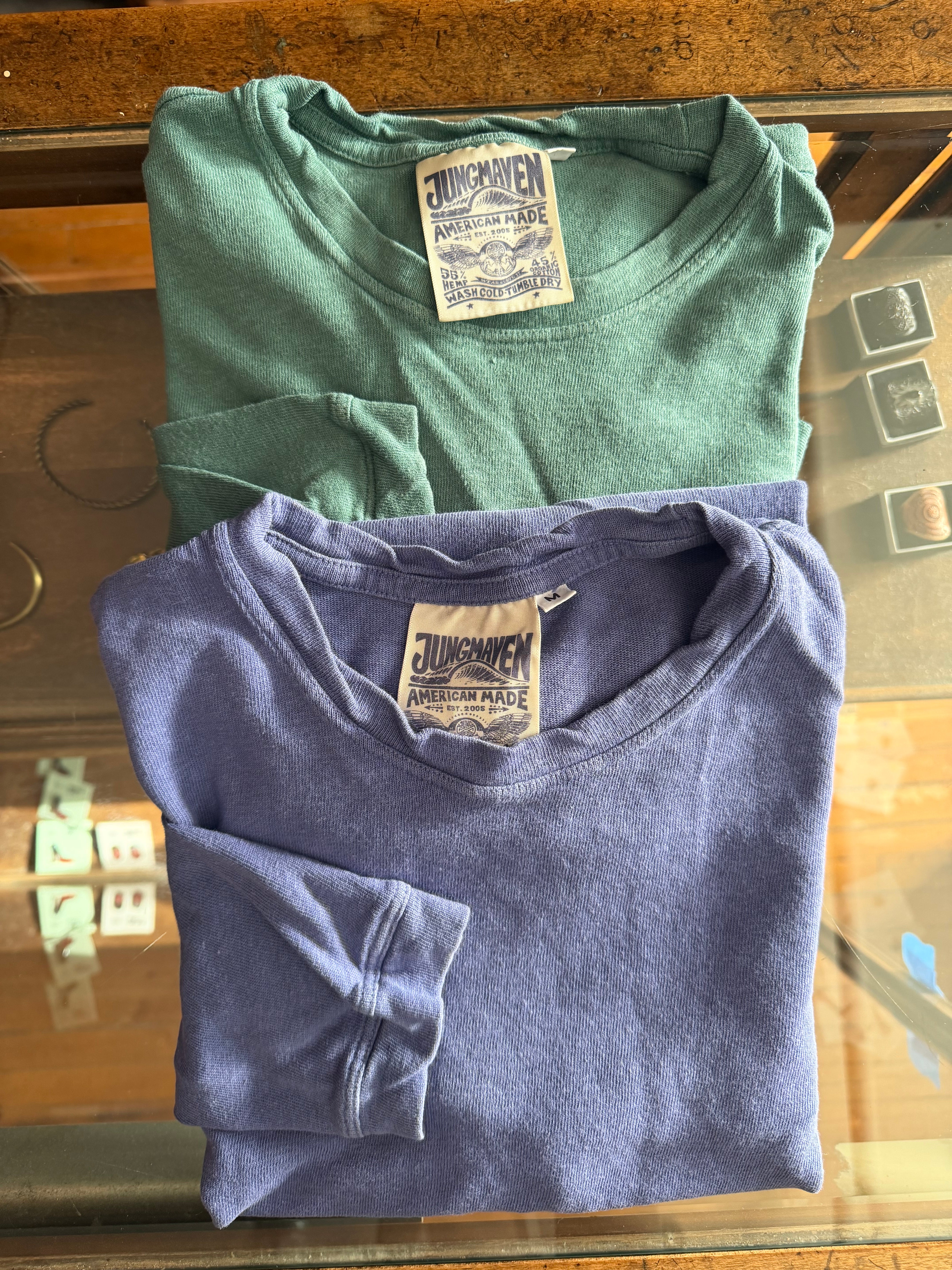 Flat lay front view of two Jungmaven crewneck tees, green and blue, showing fabric texture and tags.