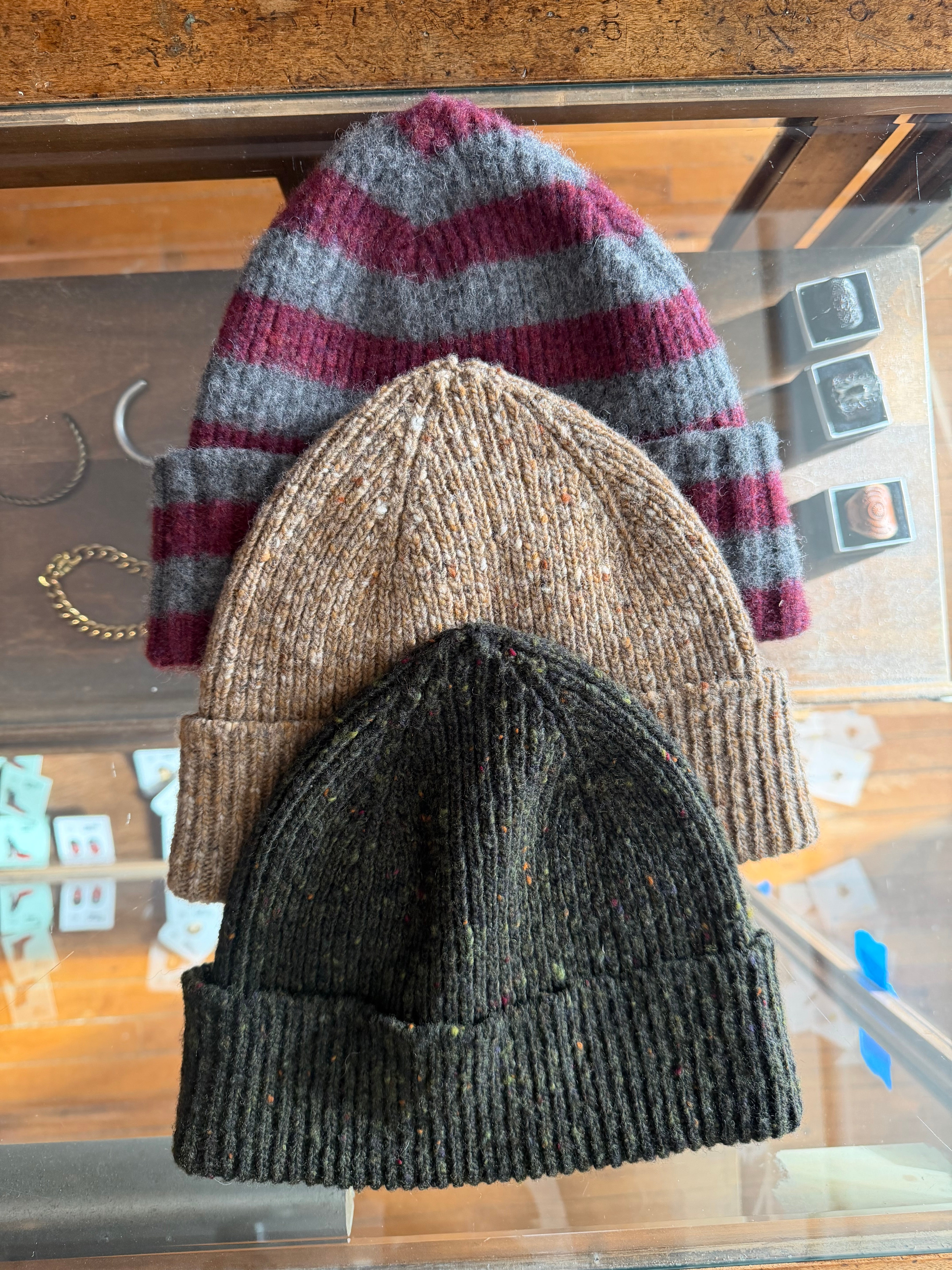 Flat lay of three knit beanies—striped grey and burgundy, speckled tan, and speckled black—displayed front side up on a glass counter at Dant shop.