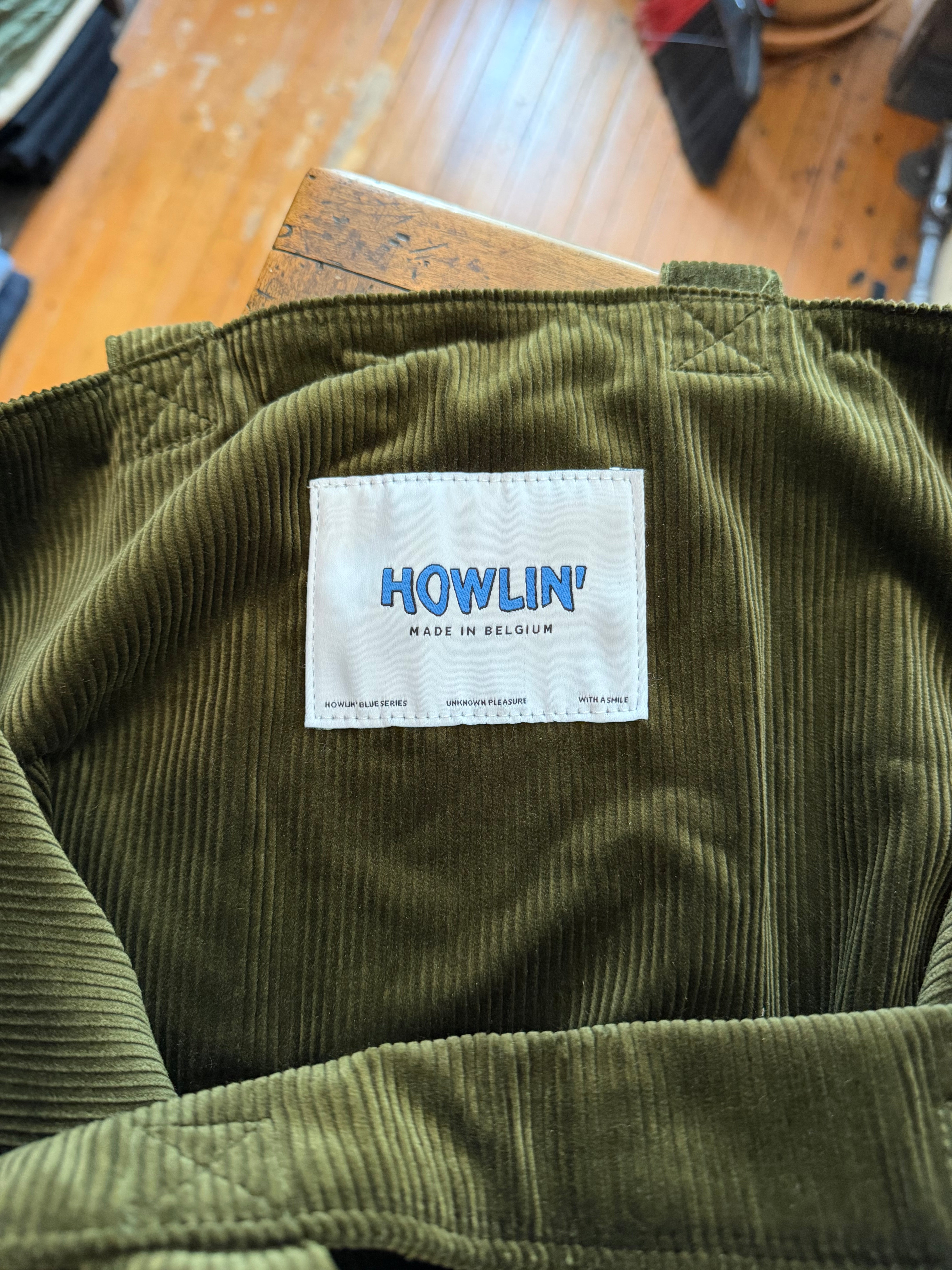 Close-up of olive corduroy garment tag, showing 'HOWLIN' Made in Belgium' label on textured fabric.