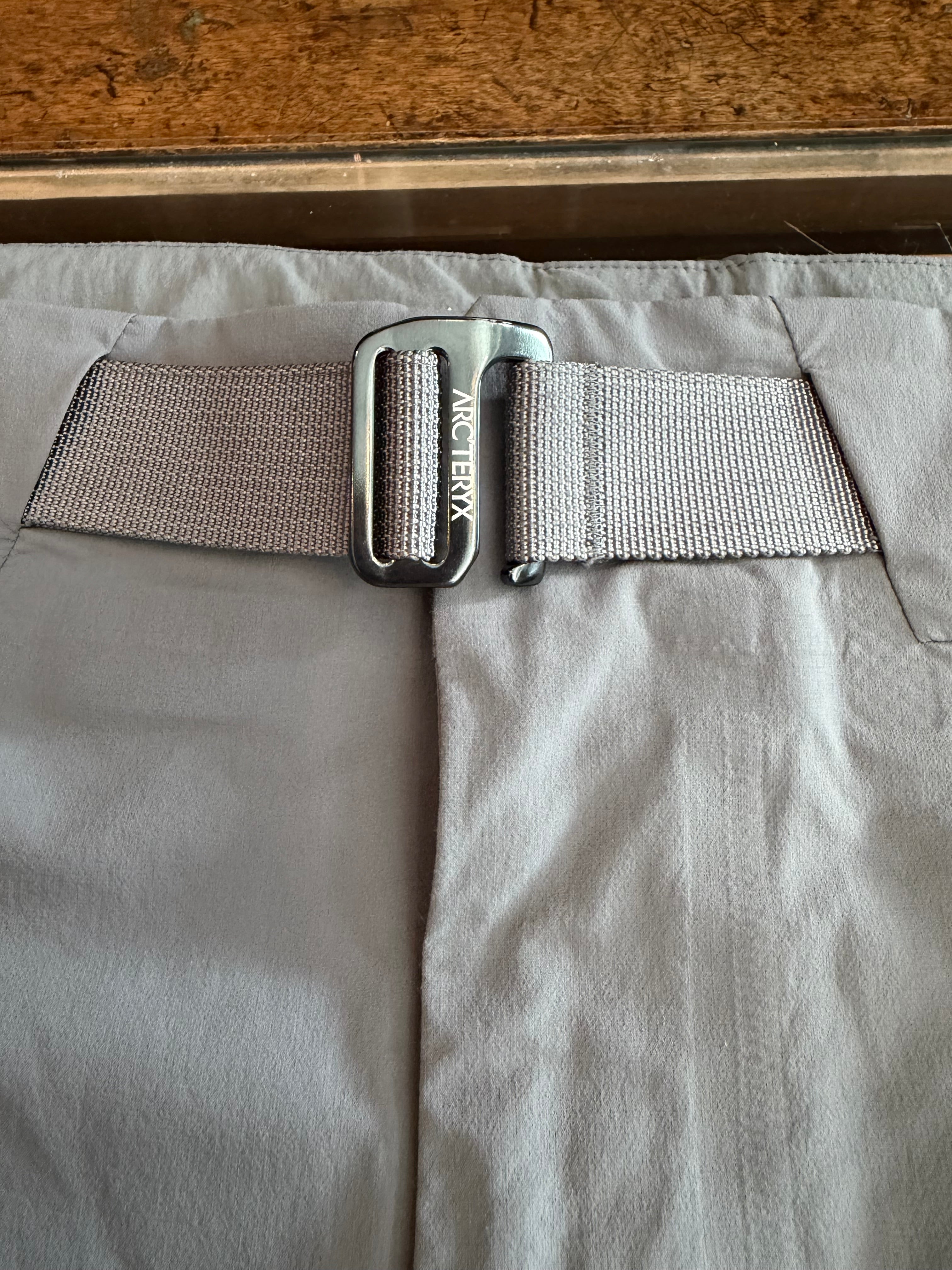 Close-up front view of gray pants with Arc'teryx web belt, metal buckle, and textured fabric waistband