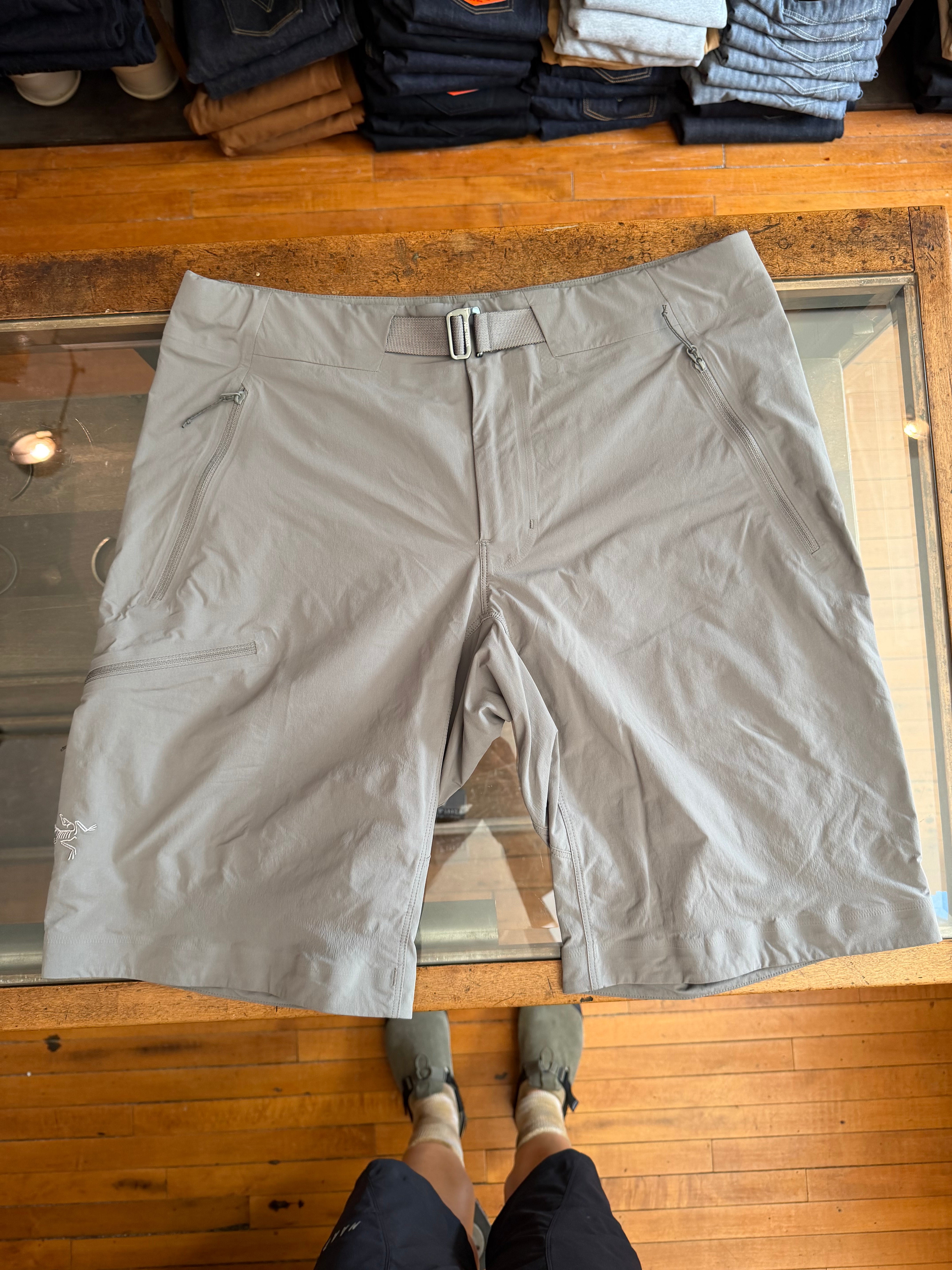 Flat lay front view of light gray men's shorts with zip pockets, belt loops, and logo on left leg.