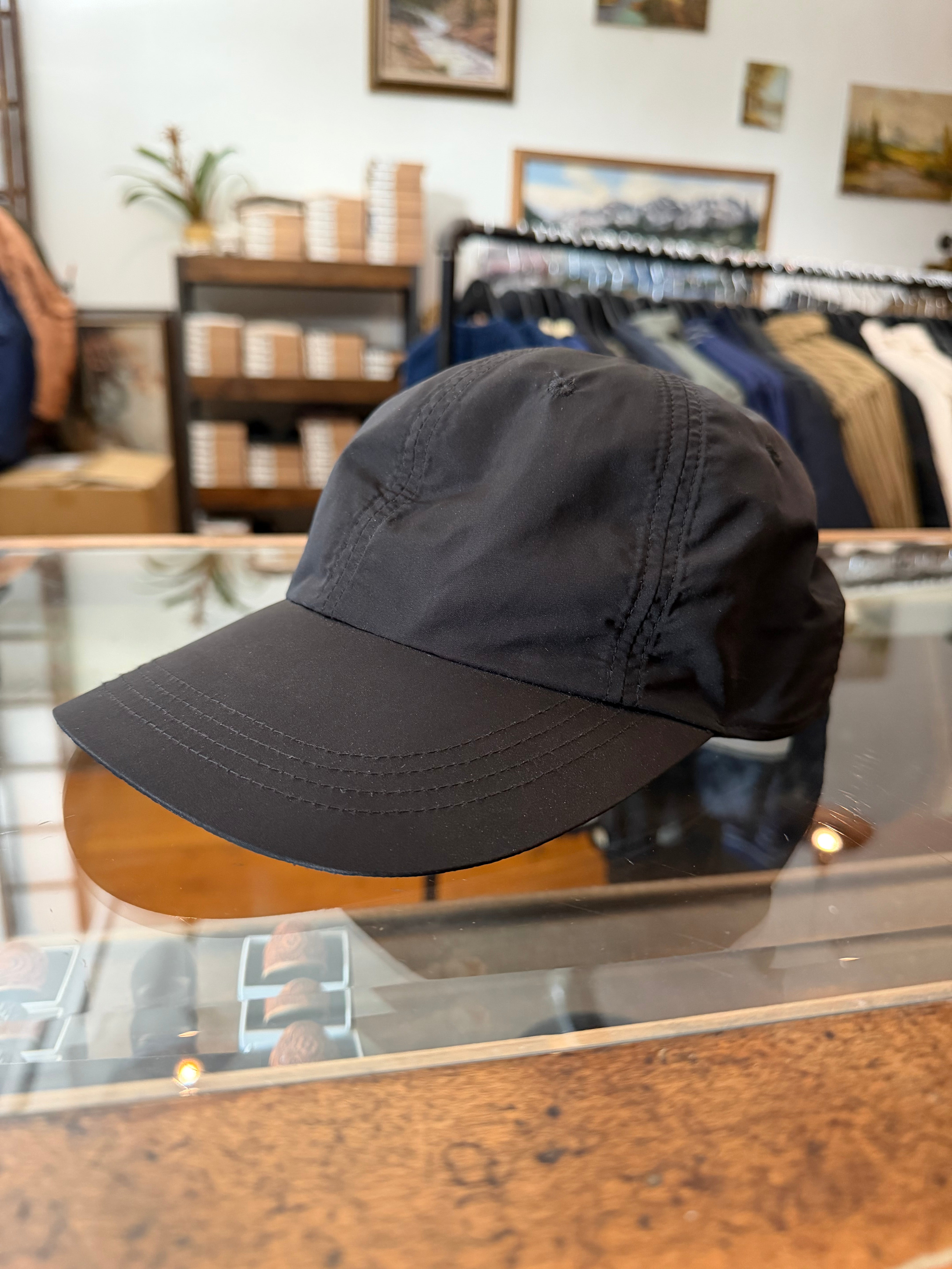 Black cap front view, showcasing brim stitching and fabric texture, displayed on glass counter in apparel store.