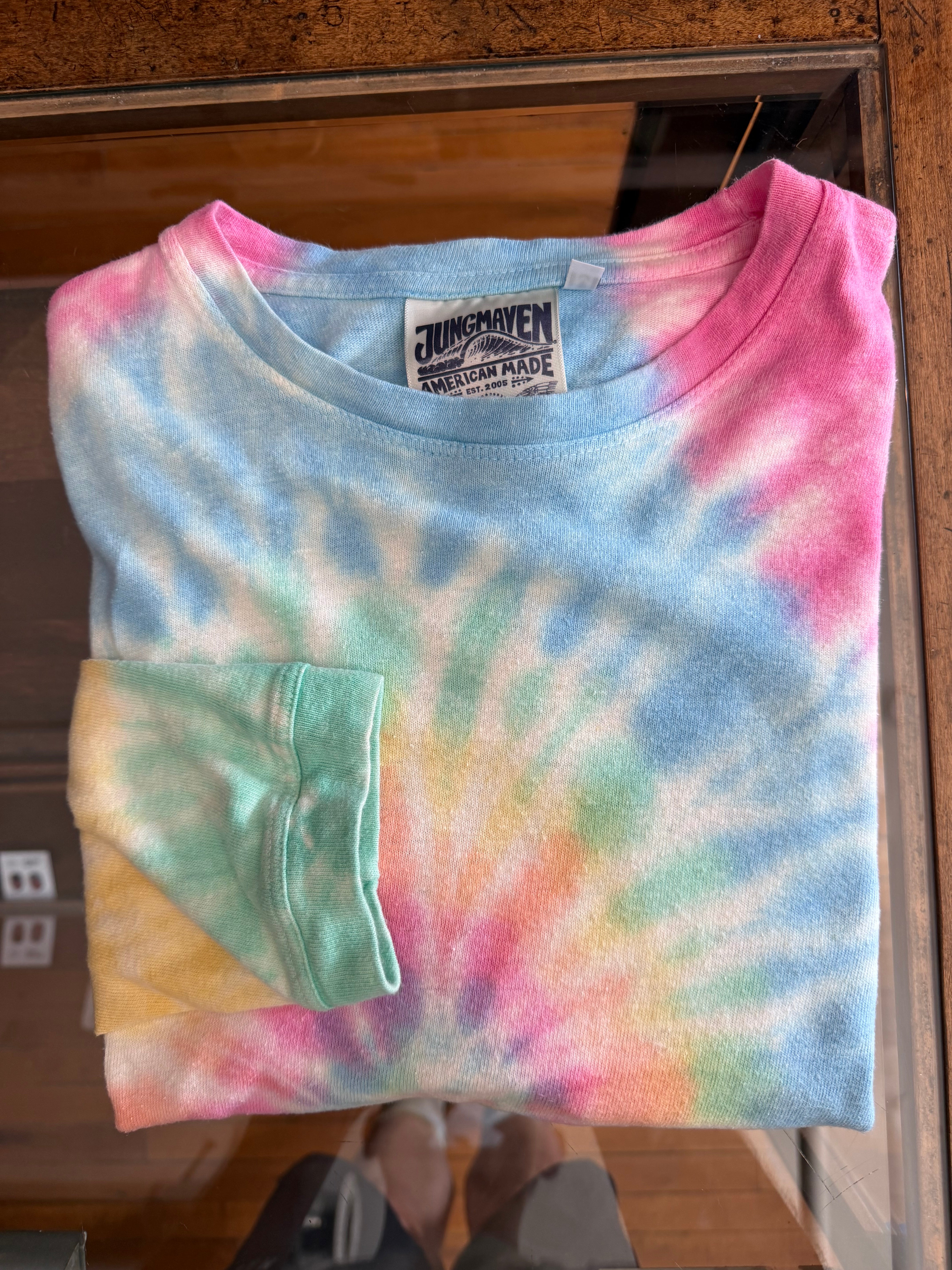 Pastel tie-dye t-shirt front view, folded, showing collar tag and sleeve hem details on wood surface