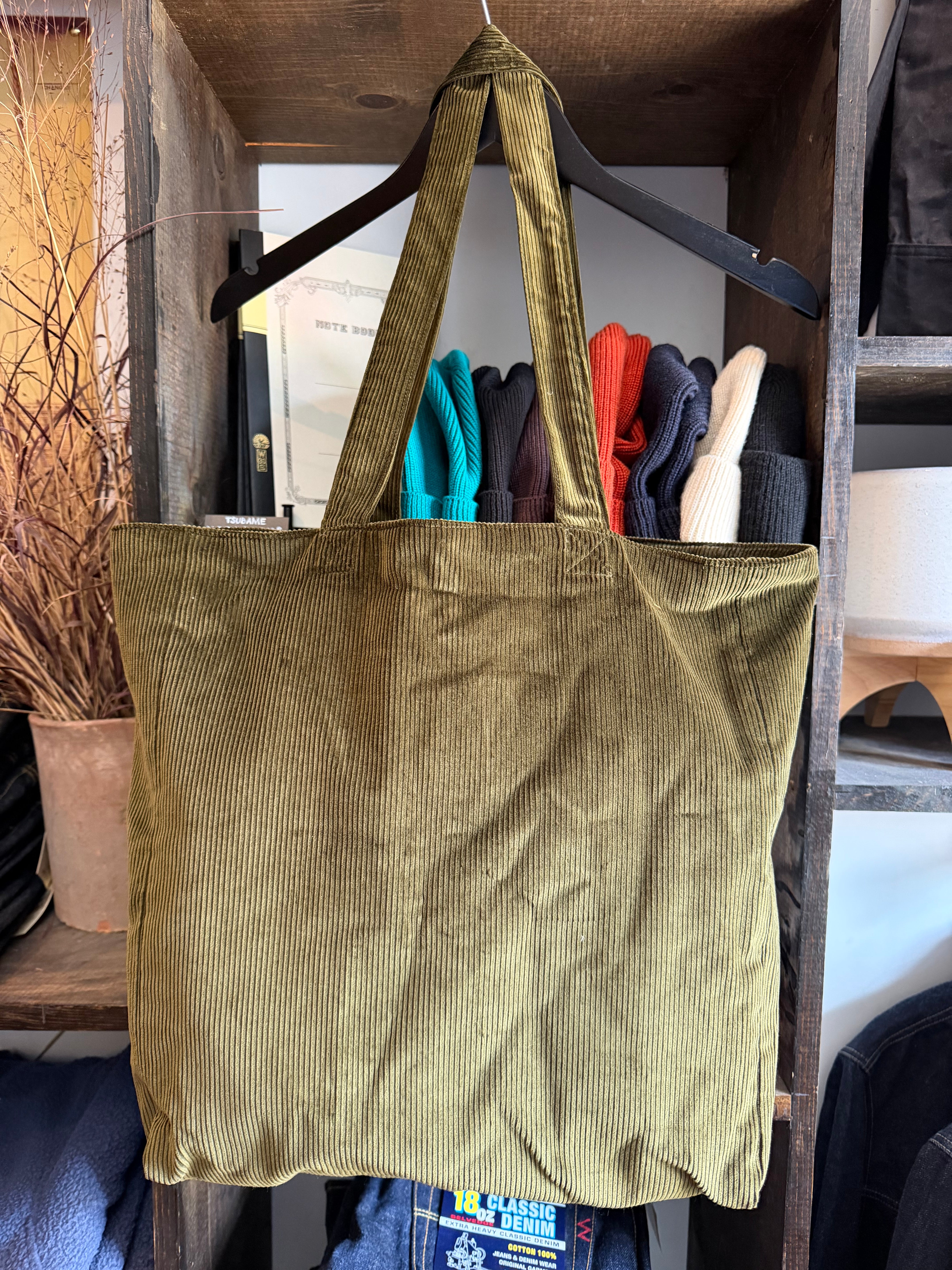 Olive corduroy tote bag front view, ribbed texture, hanging on hanger in rustic store setting