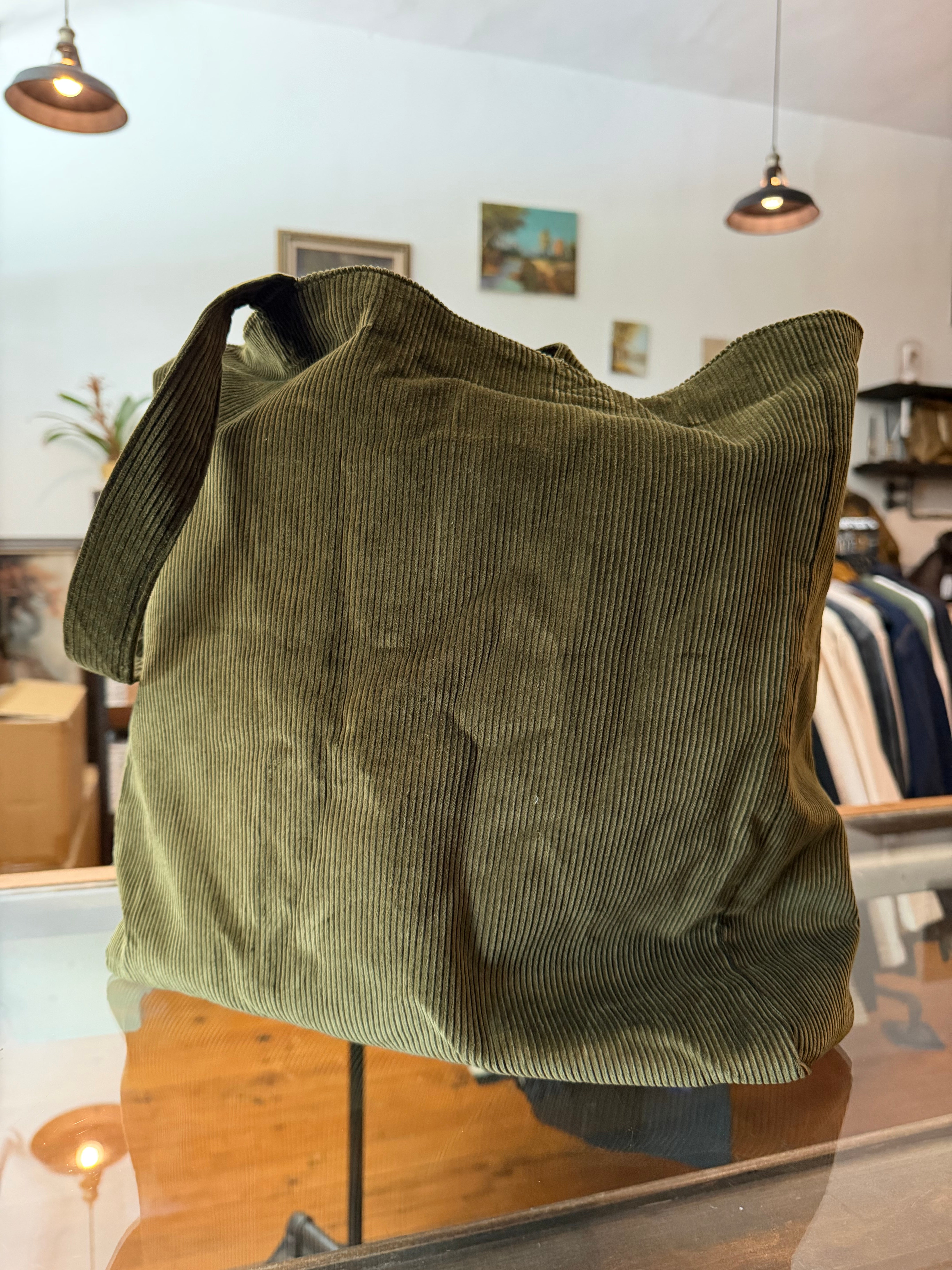 Olive corduroy tote bag front view, ribbed texture, displayed on glass counter in boutique