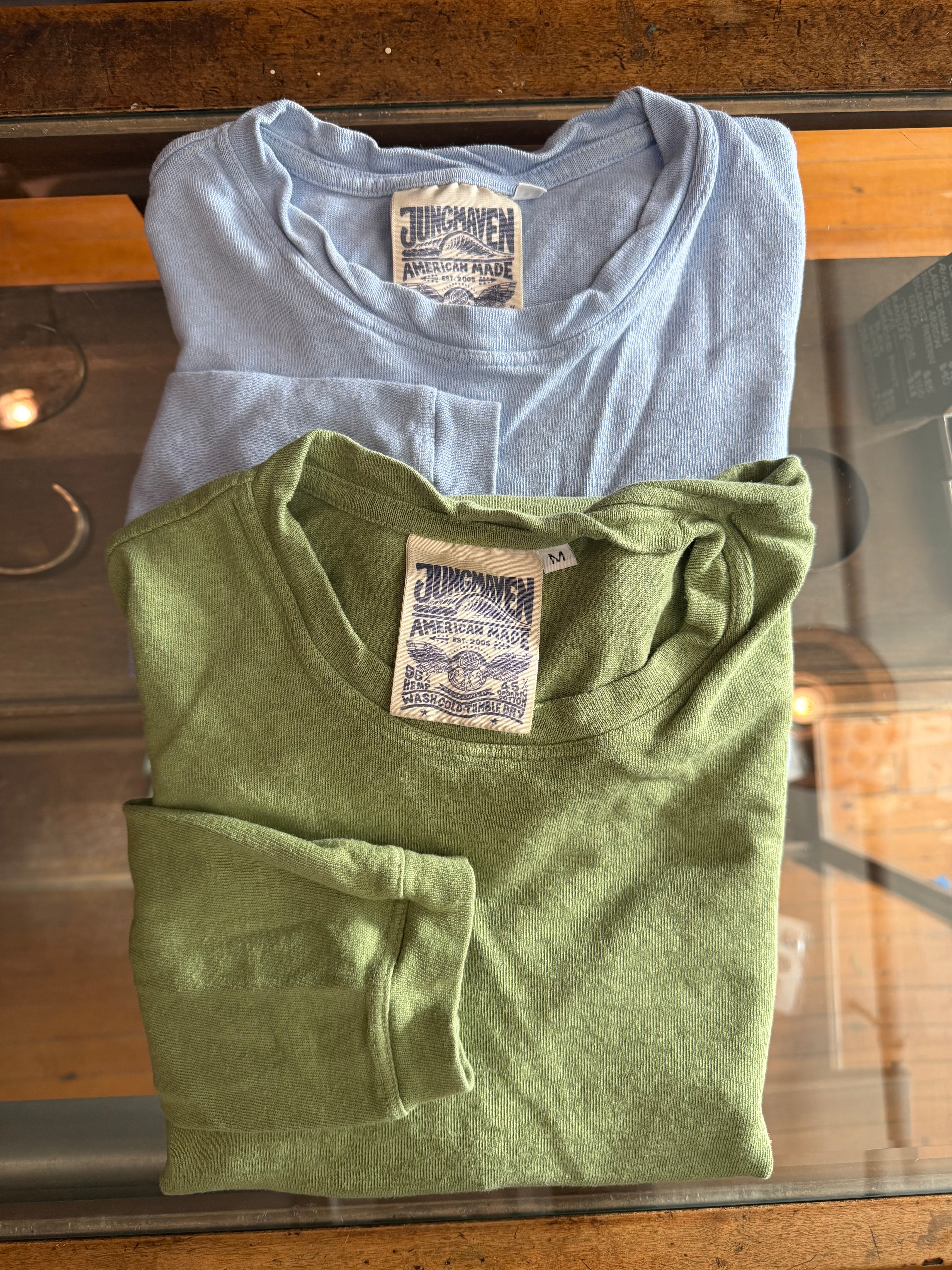 Flat lay front view of folded Jungmaven crewneck t-shirts in green and light blue, showing tags and soft fabric texture.