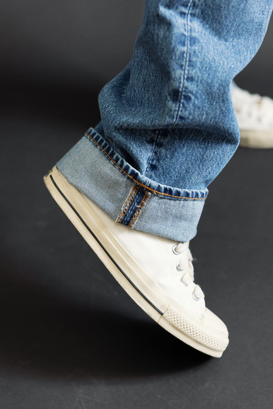 Close-up of a male model's lower leg wearing cuffed blue selvedge denim jeans and white canvas sneakers.