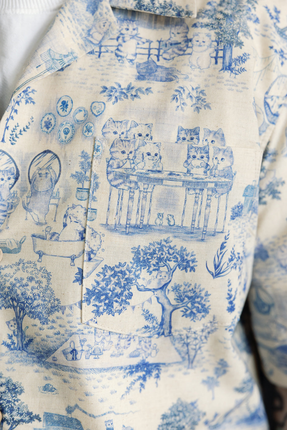 Close-up of male model's chest area wearing linen aloha shirt, front view, showing blue cat print, chest pocket, and button details. Model is 5'9