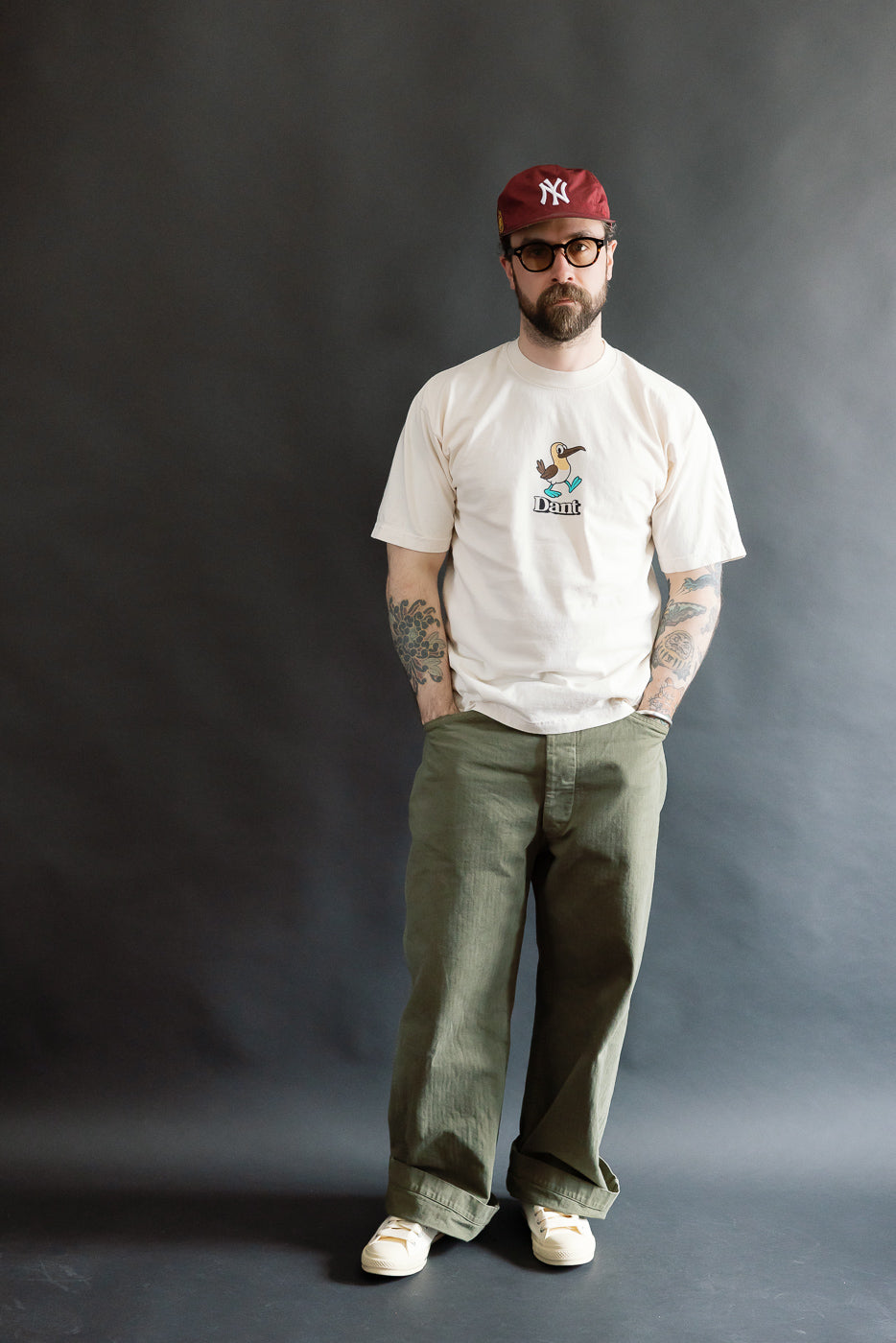 Model wearing the 6.5oz - Dant Blue Footed Booby T-Shirt - Ecru, featuring a cartoon bird graphic above Dant, paired with olive-green trousers and off-white sneakers for a casual style.