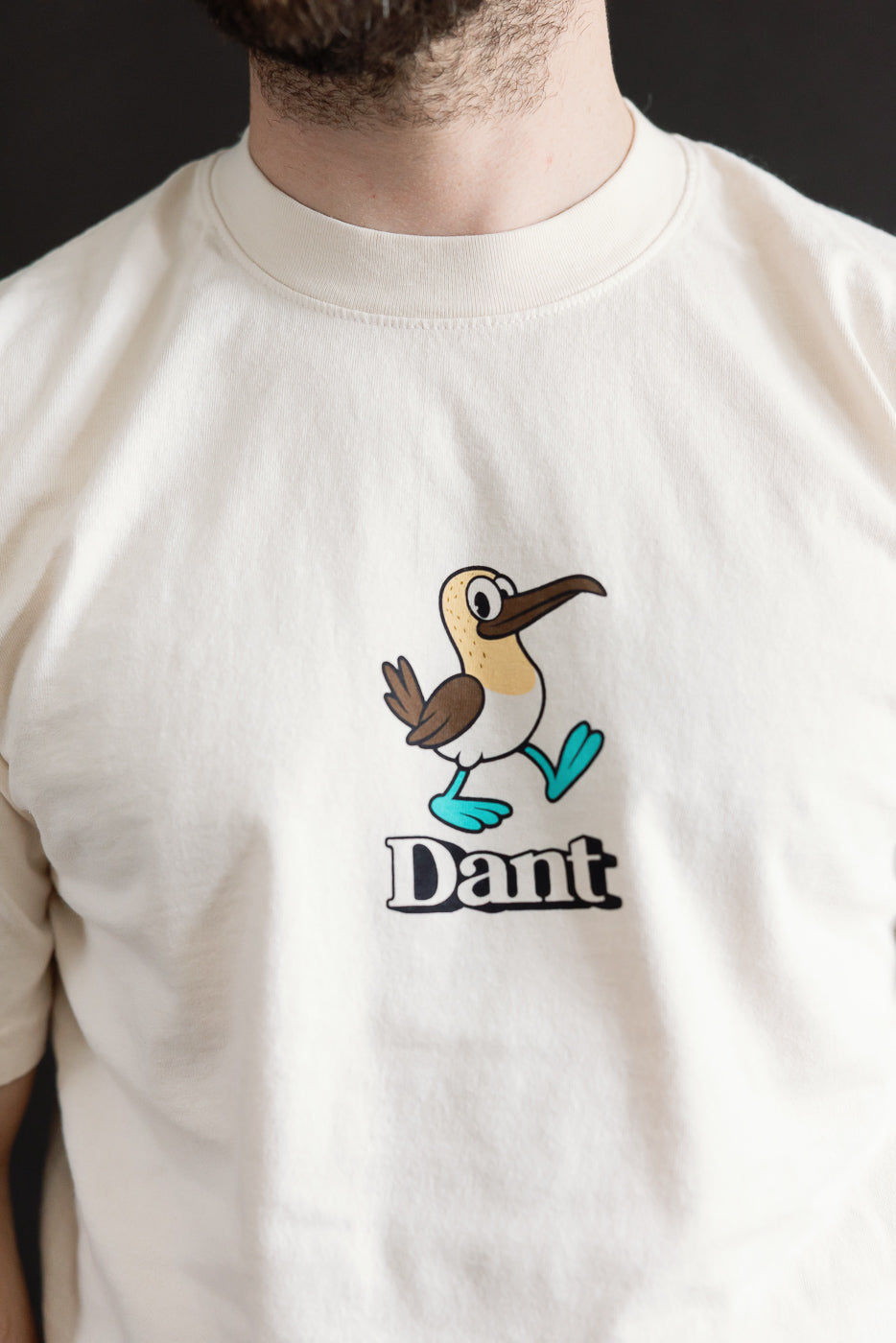 Dant Blue Footed Booby T-Shirt - Ecru features a cartoon bird with brown wings and teal feet, striding right above bold Dant lettering, embodying playful and heritage-inspired design.