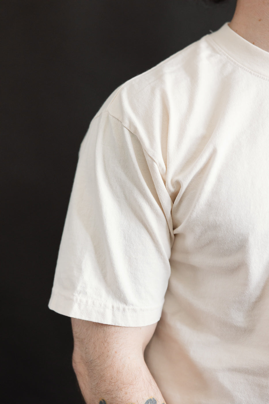 Close-up of a person wearing the 6.5oz - Dant Blue Footed Booby T-Shirt - Ecru, showcasing the sleeve seam, fabric fold, and part of a tattooed forearm, reflecting Dant's heritage-inspired style.