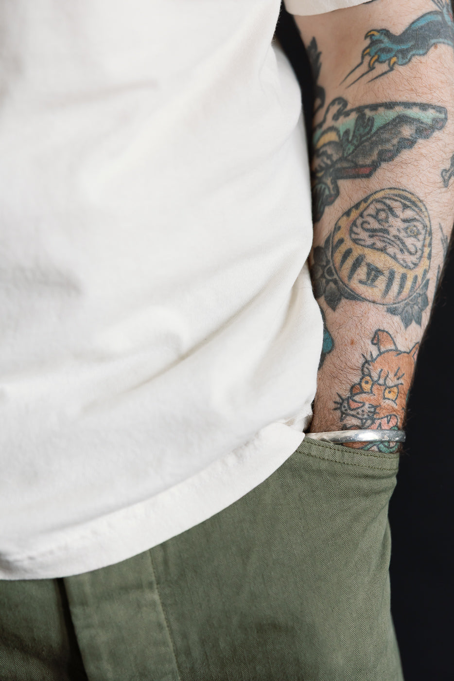 A close-up of a 6.5oz Dant Blue Footed Booby T-Shirt in ecru, tucked into olive-green pants. A silver bangle adorns the tattooed arm, enhancing the shirt's playful, heritage-inspired design.
