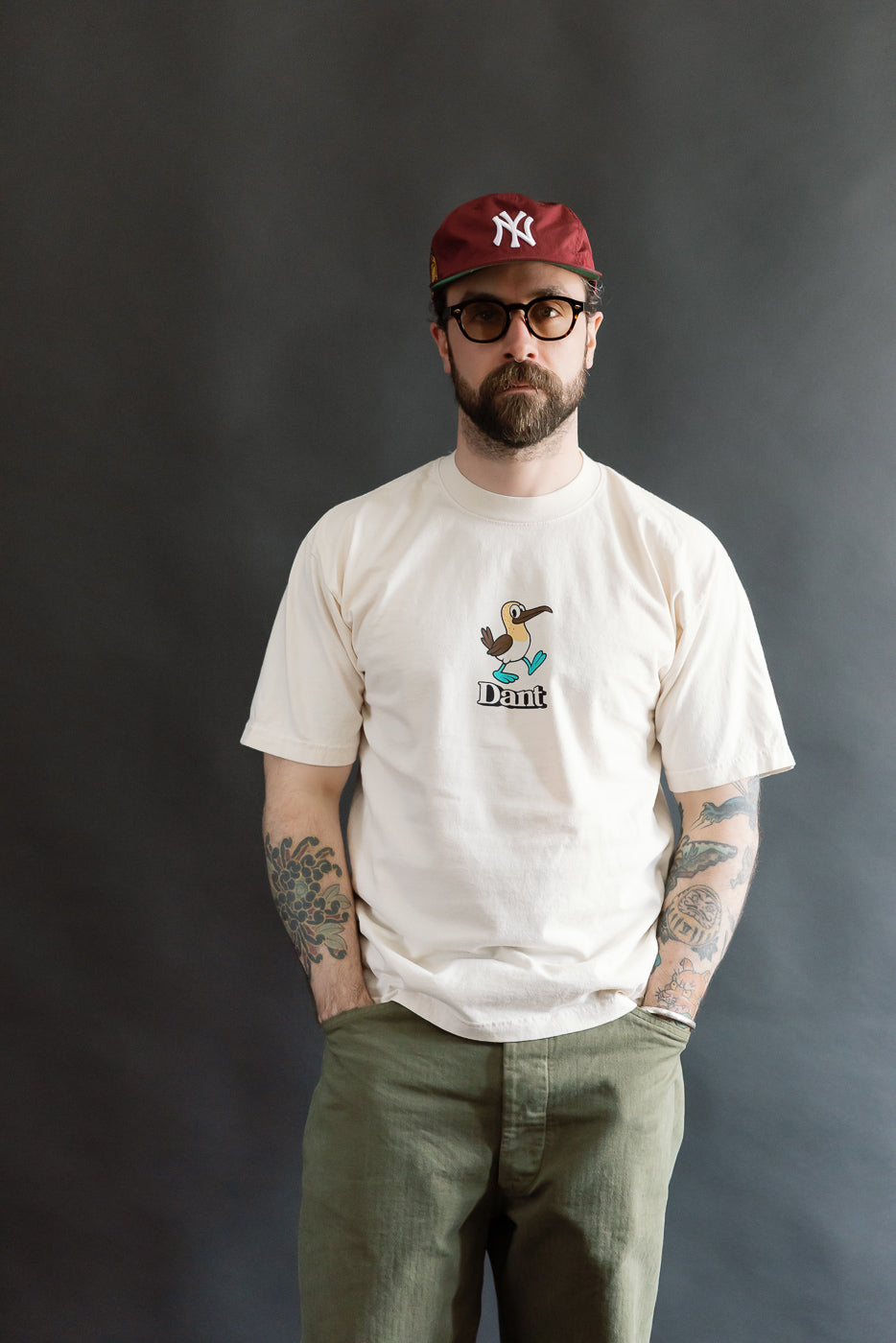 Man wears the 6.5oz Dant Blue-footed Booby T-Shirt in ecru, showcasing a cartoon bird design above Dant, paired with olive trousers and a burgundy cap, embodying relaxed, heritage-inspired style.