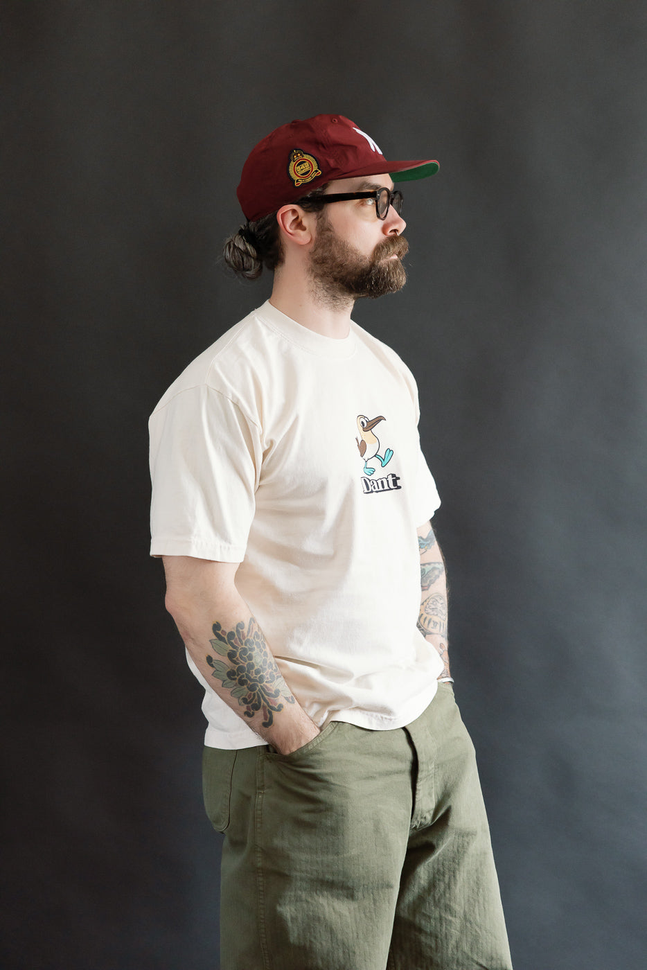 6.5oz - Dant Blue Footed Booby T-Shirt in Ecru: Modeled by a bearded man in a burgundy cap, showcasing its playful bird graphic and relaxed, oversized fit.