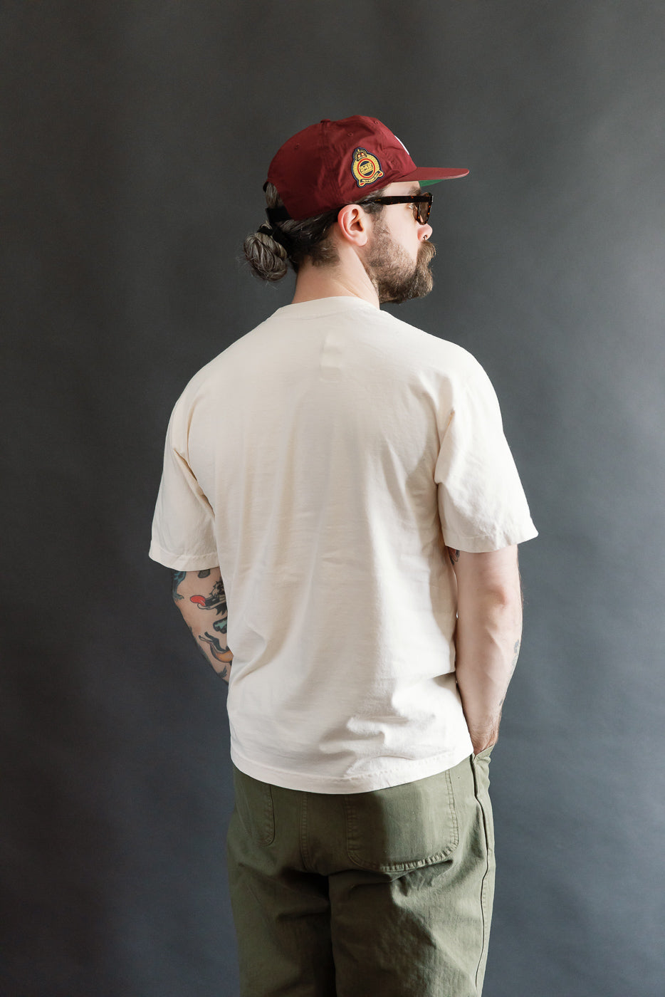 Man models the Dant 6.5oz Blue Footed Booby T-Shirt in Ecru, showcasing its oversized fit with sleeves and chest illustration. Styled with cargo pants and red cap, reflecting Dant's heritage-inspired fashion.