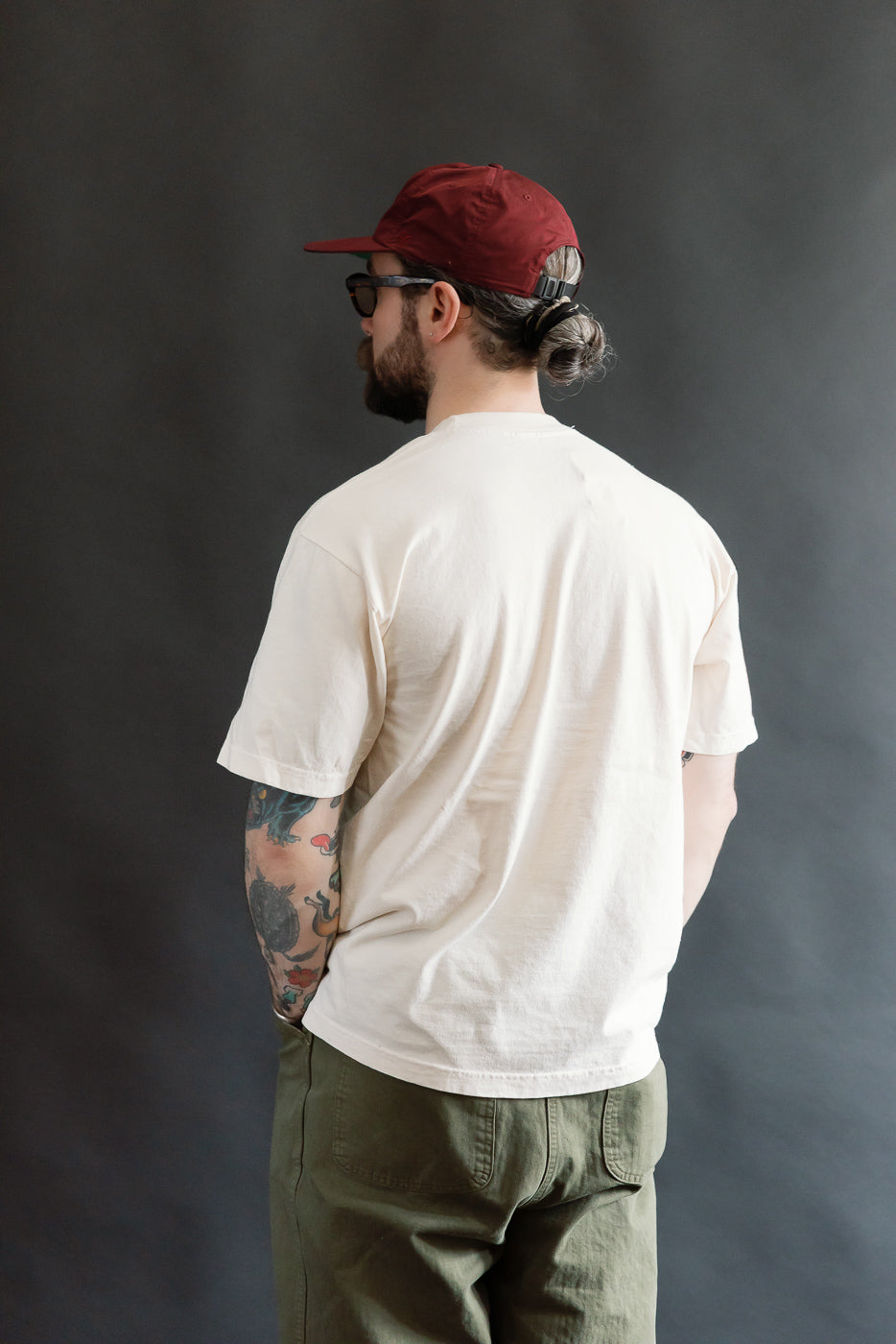 Man wearing the 6.5oz - Dant Blue Footed Booby T-Shirt - Ecru, featuring a thick ribbed collar and colorful tattooed forearm, styled with olive-green pants and a burgundy baseball cap.