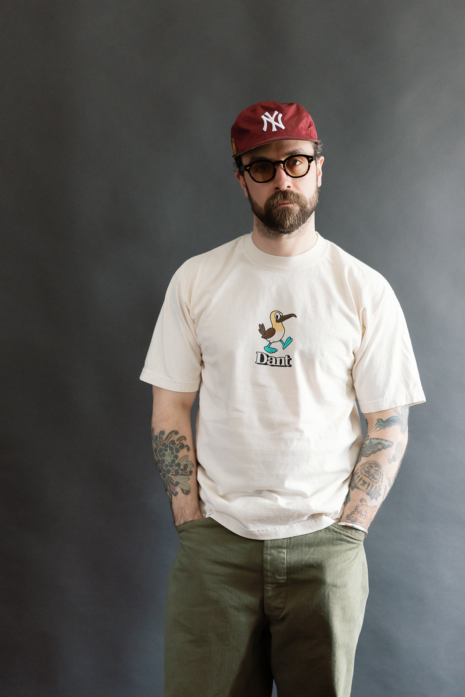 Model wearing a 6.5oz Dant Blue Footed Booby T-Shirt in Ecru, featuring a cartoon bird print and Dant text, paired with olive-green pants, showcasing a modern, boxy fit.
