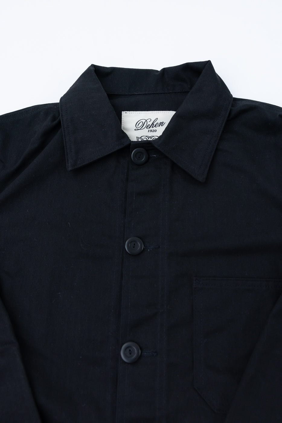 Close-up of front upper section of black dry wax chore coat showing buttons, collar, chest pocket, and collar tag.