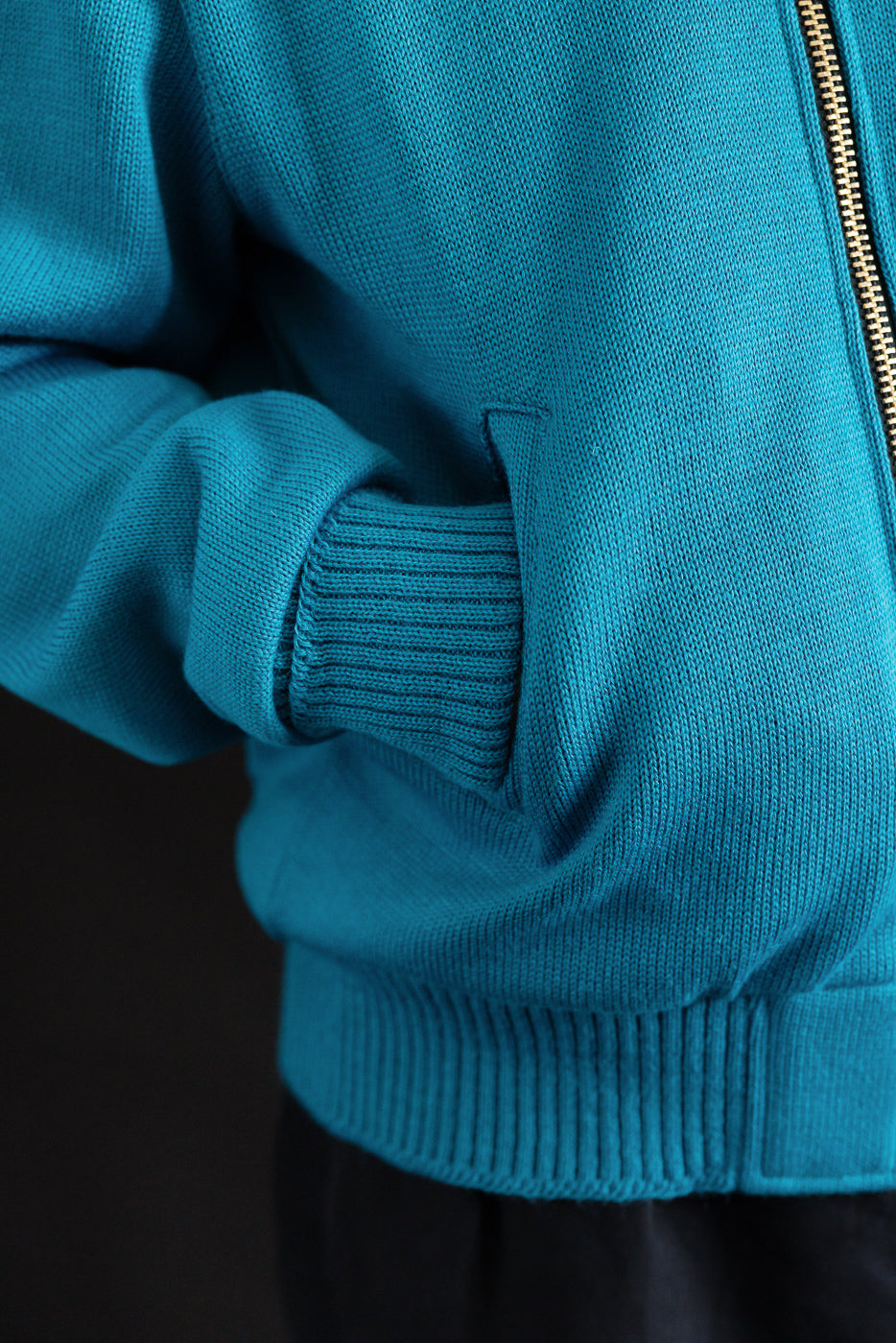 Close-up of male model's hand in front pocket of aqua full zip moto hoodie, showing ribbed cuff and textured knit fabric.