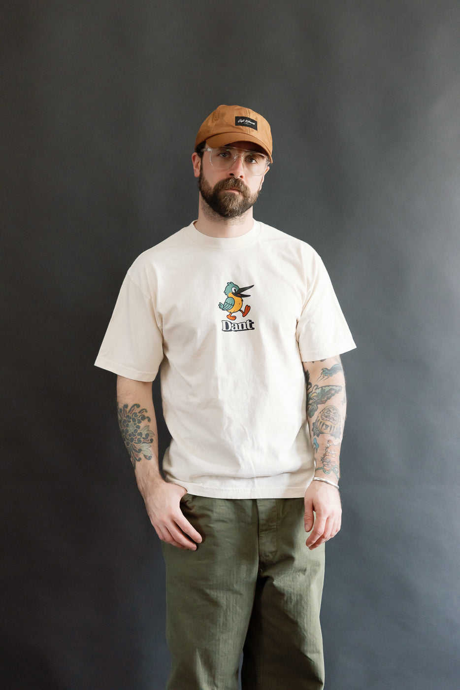 A man models the 6.5oz - Dant Kingfisher T-Shirt - Ecru, featuring a cartoon bird graphic. He pairs it with olive-green pants and a tan cap, embodying Dant's heritage-inspired style.