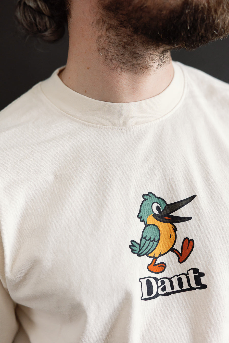 Dant 'Kingfisher' T-Shirt - Ecru features a cartoon bird with teal wings and orange body. Bold 'Dant' text underneath. The off-white shirt has an oversized fit with a thick ribbed collar.